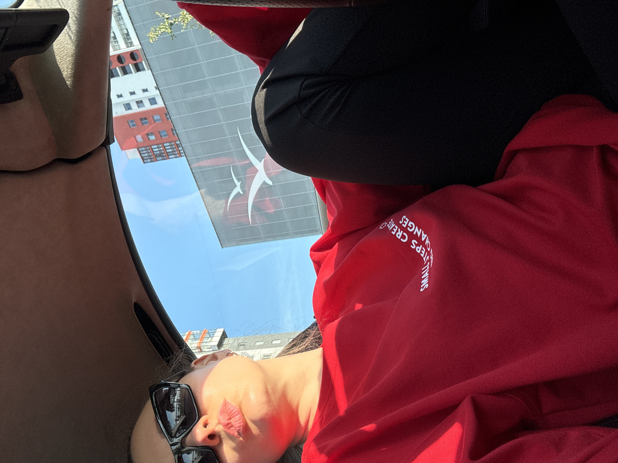The image depicts a rotated, bright daytime scene captured from inside a vehicle in what is identified as Saint Petersburg, Russia. The perspective shows a partial view of a person and the exterior world through a car window.
The primary subject is a person, visible from the mid-torso upwards, angled as if leaning back or looking up. They are wearing a red long-sleeved top, possibly a sweatshirt or hoodie, with white text on the right shoulder/chest area that reads, "SMALL STEPS CREATE CHANGES." The person's face is partially visible, with dark sunglasses resting on their nose, and their lips are slightly parted, showing a pinkish hue. Dark hair is also discernible. In the upper right of the frame, a segment of black clothing, likely leggings or trousers, is visible.
The interior of the vehicle, characterized by light beige fabric (possibly a headliner or pillar), frames the left side of the image. Through the vehicle's window, a clear blue sky and several multi-story buildings are visible. One prominent building features distinct red and white sections with many windows, while others appear darker and more industrial. A white decal, resembling two stylized birds or abstract flying shapes, is affixed to the car window, overlaying the external view.
The scene suggests transit through an urban environment on a sunny day.