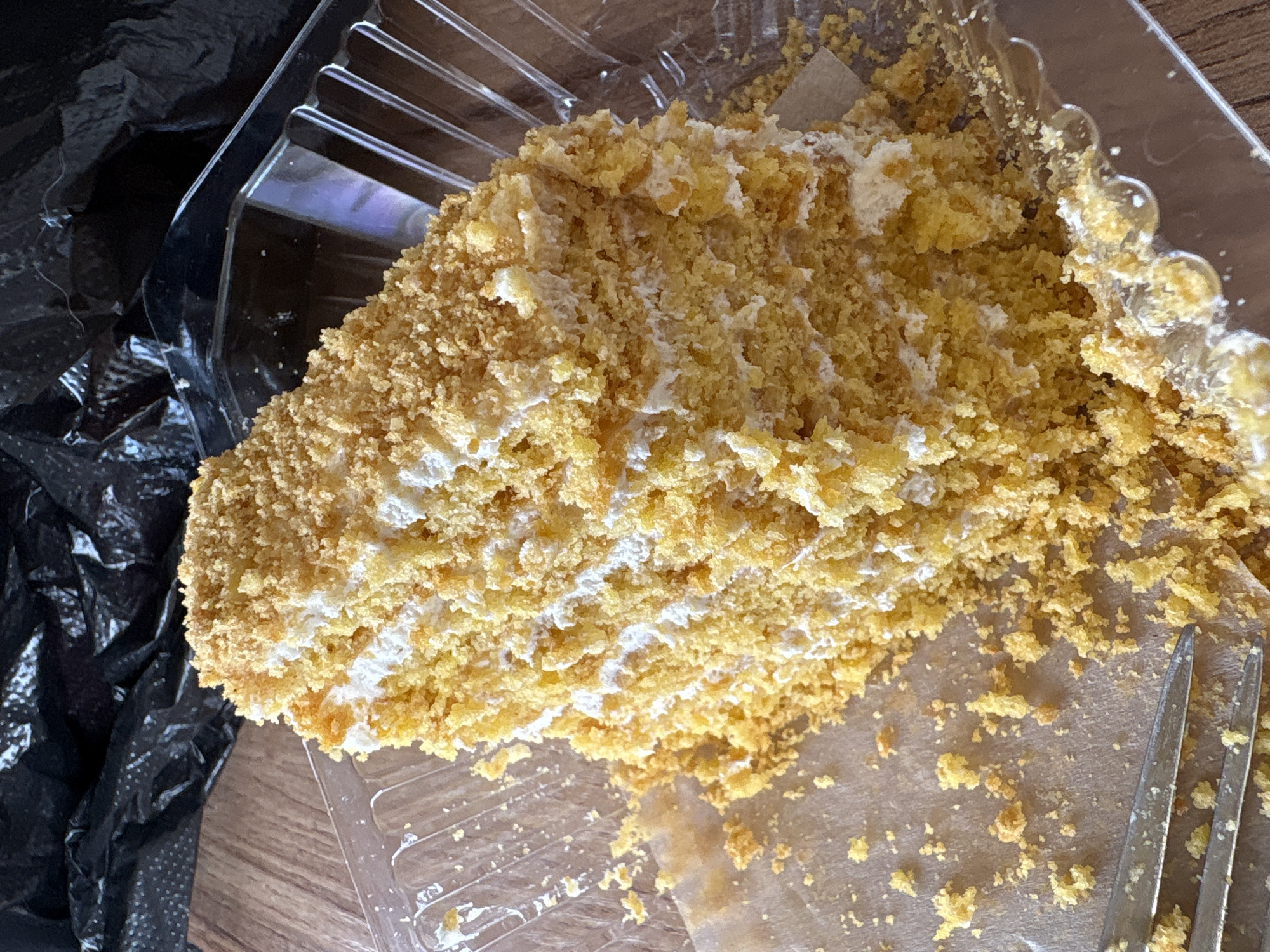 A piece of cake rests in a clear plastic container. Crumbs are scattered around the cake on the container's surface. A black plastic bag is visible in the upper-left corner, partially obscuring the container. The container is on a wooden surface. Two metal forks are in the container next to the cake. The location is Ембаево, Russia.