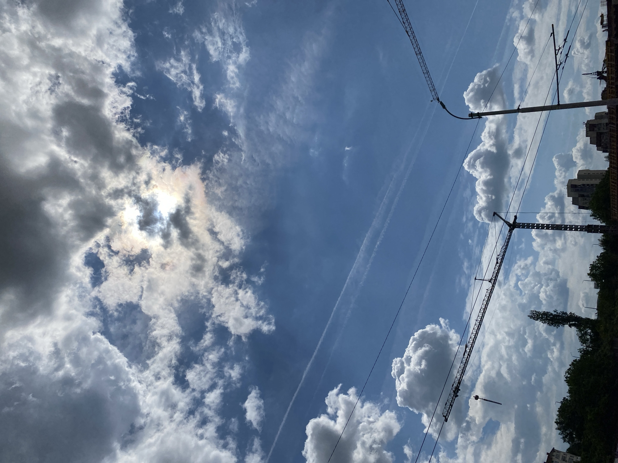 The image depicts a daytime scene in Pleven, Bulgaria. The sky is predominantly blue with scattered cumulus clouds. The sun is partially obscured by clouds in the left side of the frame, creating a diffused light effect. Two contrails are visible stretching across the sky. Tower cranes are present on the right side of the frame, silhouetted against the sky. Buildings and trees are visible on the right, situated in the background. Overhead power lines are also visible against the sky.