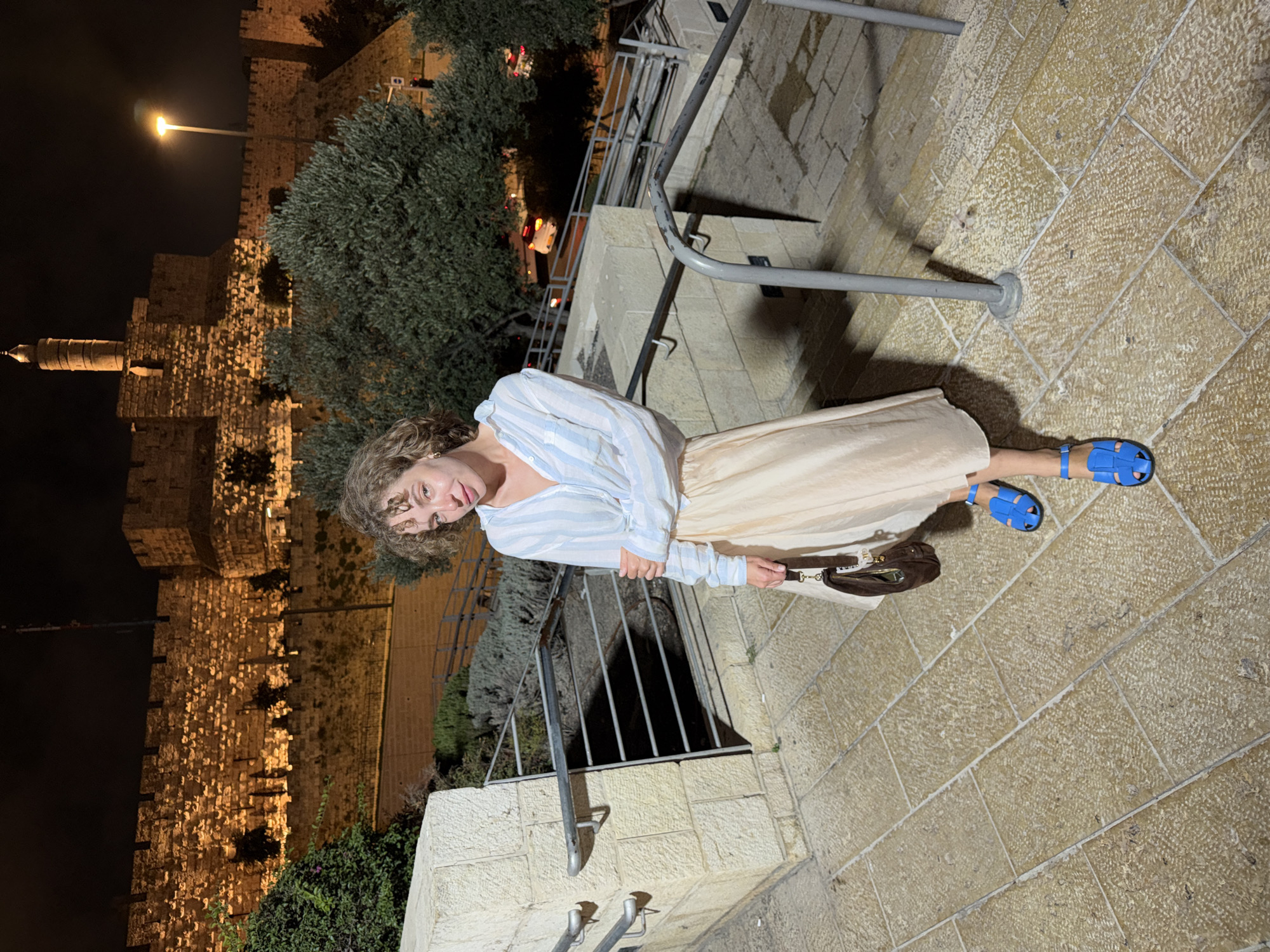 A woman stands on a stone staircase at night in Jerusalem, Israel, with a historic stone wall illuminated behind her. She is wearing a light blue and white striped button-down shirt tied at the waist over a long, beige satin skirt. Her feet are clad in bright blue sandals. In her right hand, she carries a small, dark brown purse with a gold clasp. The staircase is made of large, textured stone pavers, and a metal railing runs alongside it. The ancient stone wall in the background, likely part of the Old City fortifications, is lit by artificial lights, casting a warm glow on the rough-hewn stones. Trees and greenery are visible in front of the wall, and a street with car lights can be seen in the distance. The scene suggests an evening outing in a historically significant area of the city.