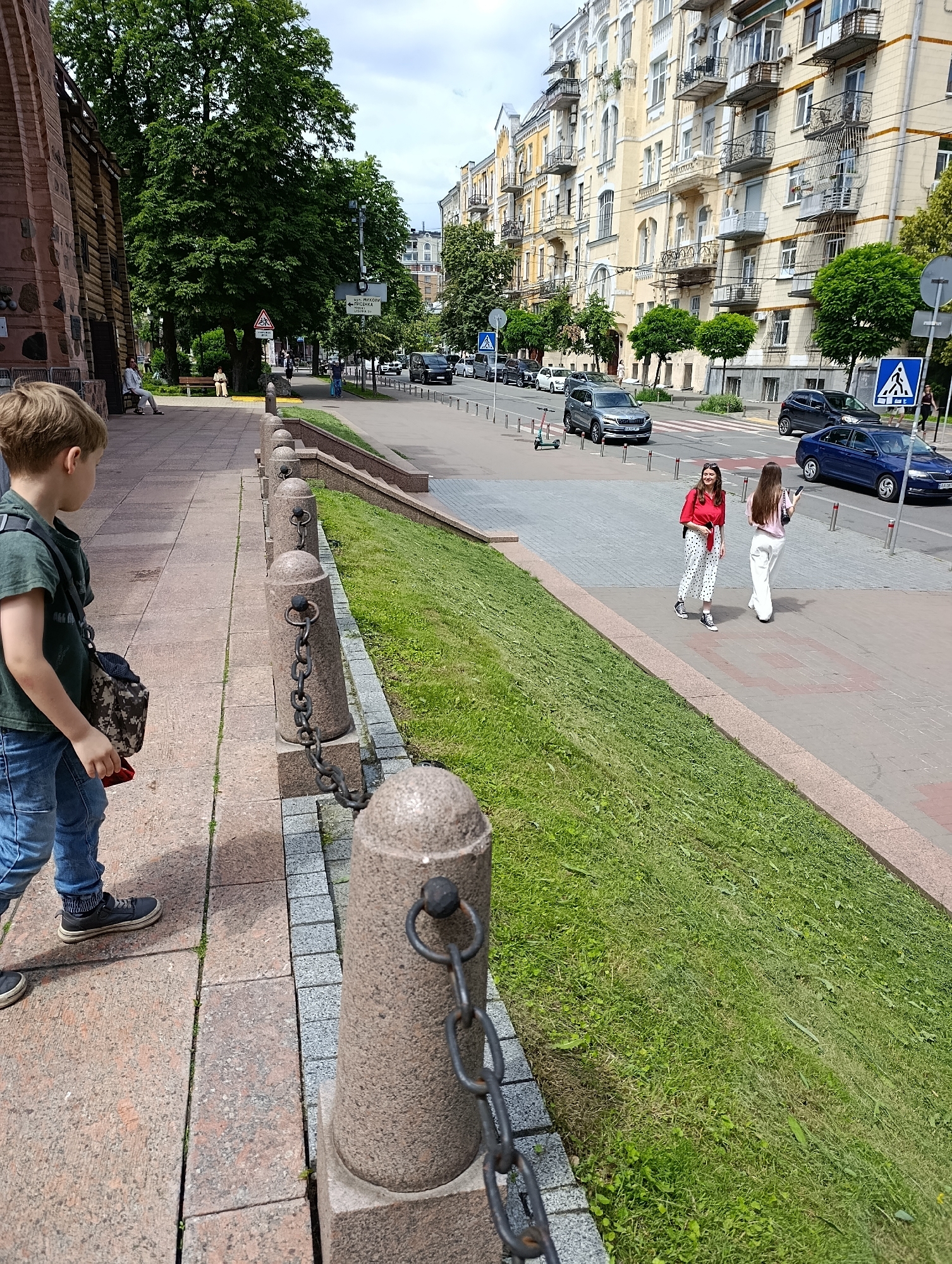 This is a street-level, medium shot of a sidewalk with a boy in the foreground looking to the left. The boy is wearing a green t-shirt and blue jeans and has a camouflage bag slung over his shoulder. To the right of the boy, a grassy slope descends to a street. On the street, there are cars and two young women walking. The woman on the left wears a red top and white polka dot pants, and the woman on the right wears a light pink top and white pants. In the background, there are apartment buildings with balconies and trees. The scene appears to be in Kyiv, Ukraine, specifically on a sidewalk near the St. Nicholas Cathedral, given the architectural style of the buildings and the presence of a street sign with Ukrainian text.
