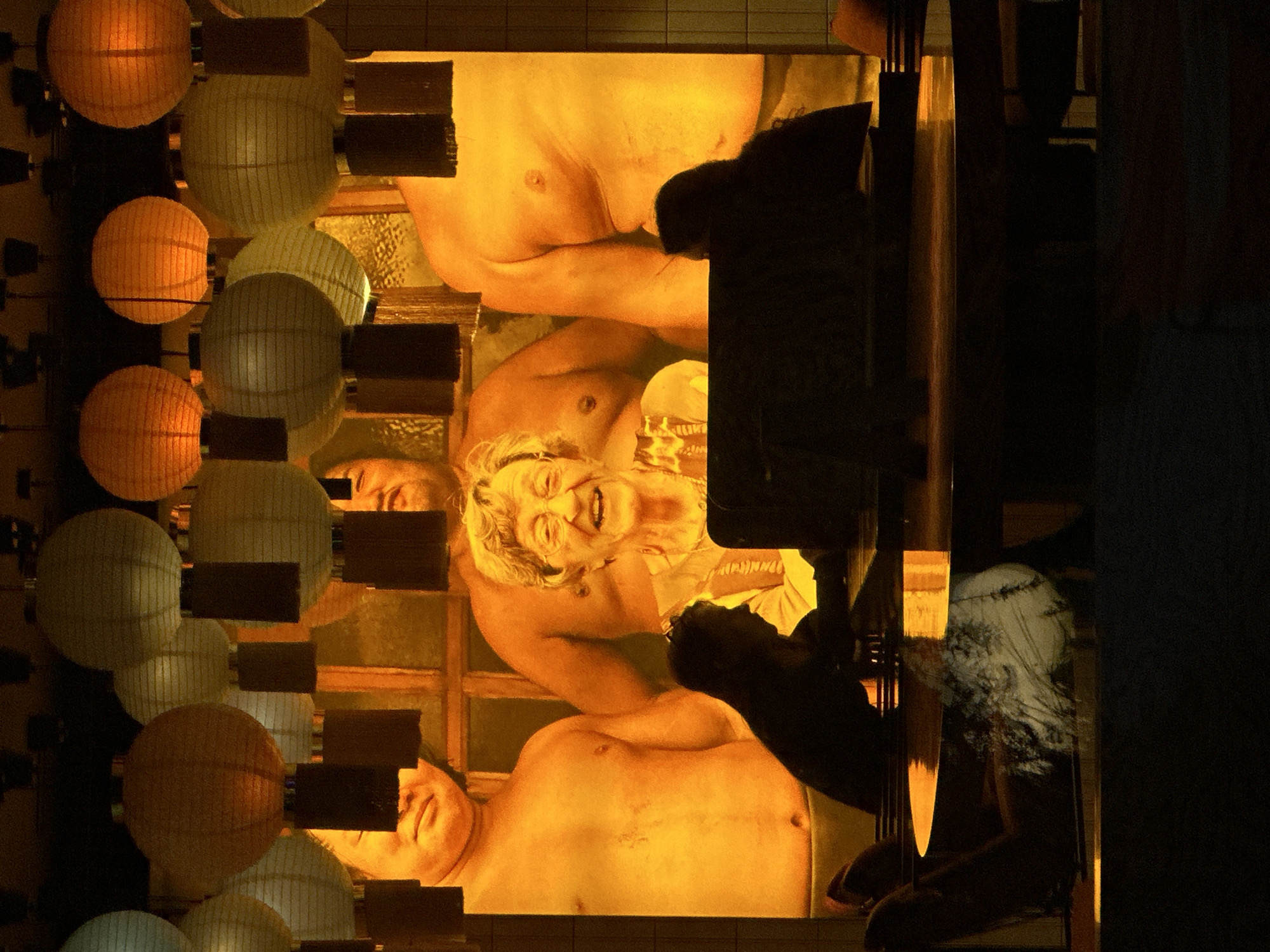 A low-angle shot shows an interior scene with lighting fixtures hanging near a window displaying a painting. On the left side of the frame, a series of round, illuminated paper lanterns are suspended, alternating between orange and white. In the center, a window pane is visible, displaying a painting of three figures against a muted yellow backdrop. The figures are depicted without clothing, with the central figure laughing. A dark object casts a shadow to the right of the painting. The scene is shrouded in low lighting, with the right side of the image transitioning into complete darkness.