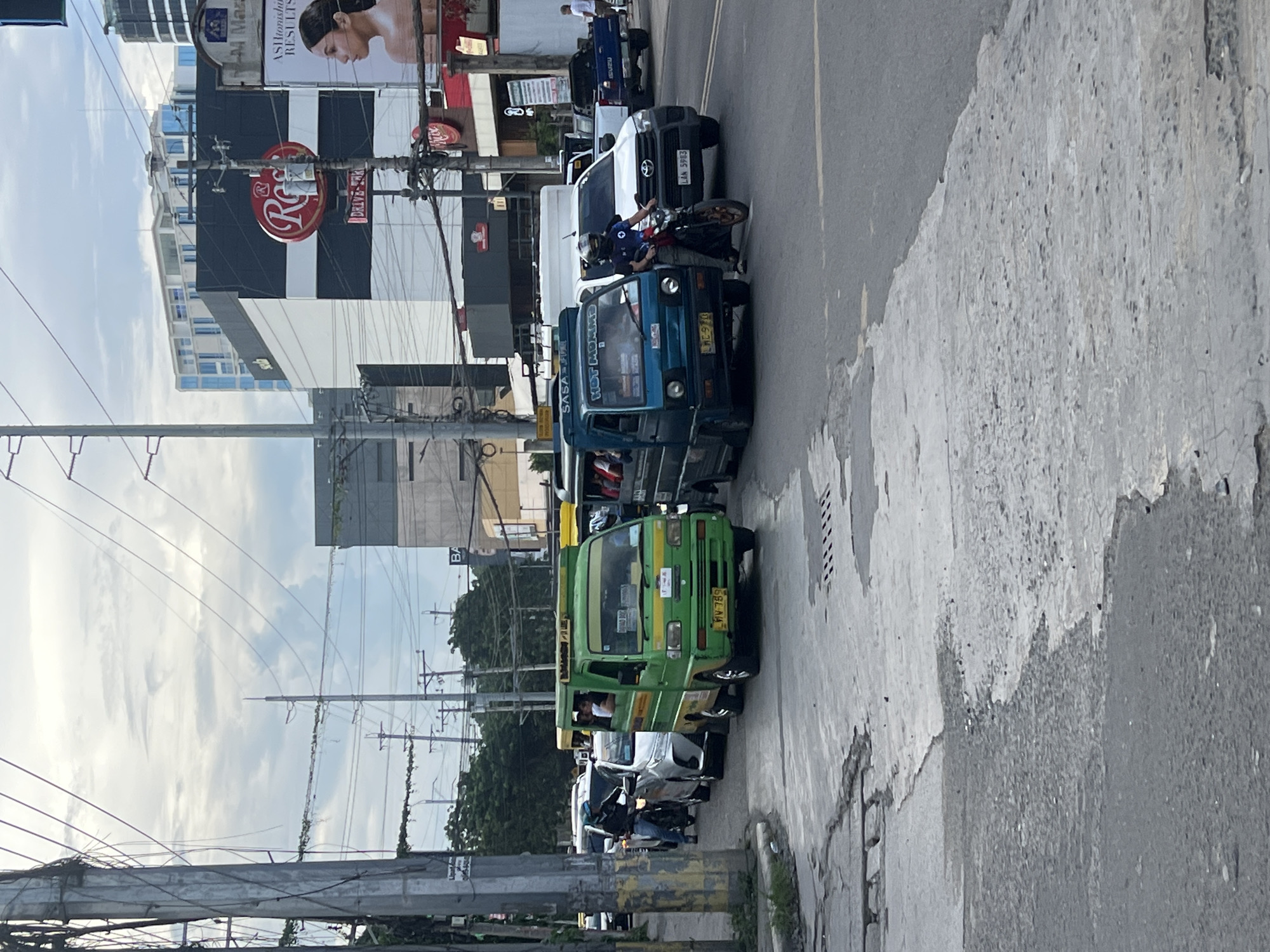 The image shows a street scene in Davao City, Philippines. A line of public utility vehicles, primarily jeepneys, is visible along the right side of the road. A motorcycle and several other vehicles are also present. A motorcycle rider appears to be conversing with someone inside one of the jeepneys. The street is flanked by buildings of varying heights, utility poles with overhead wires, and advertising signs, including one with the letter "R" and another for "AShtonishu Results". The sidewalk is cracked and shows signs of wear.