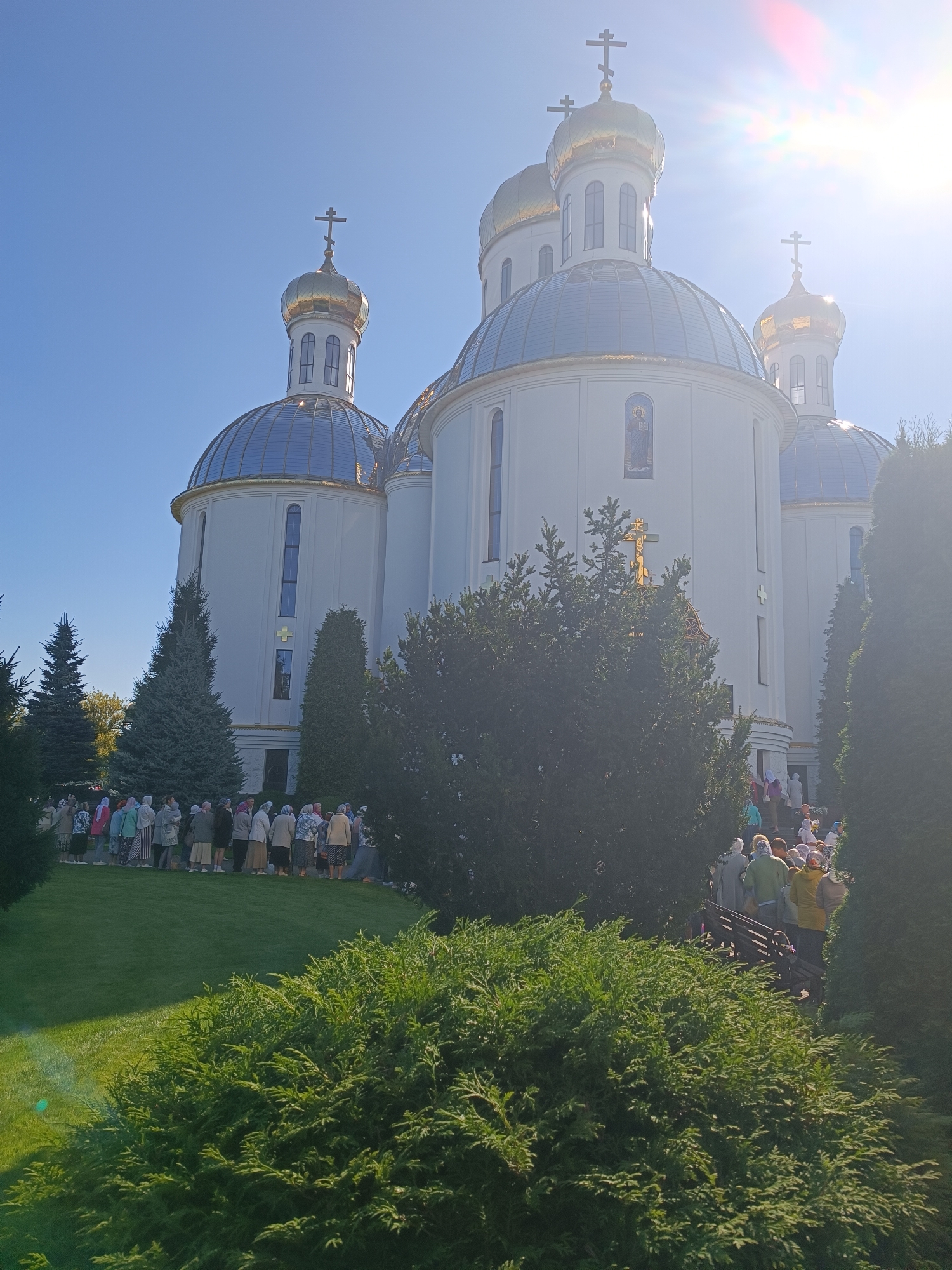 The image shows the Holy Resurrection Cathedral in Brest, Belarus on a sunny day. A group of people, mostly women wearing headscarves, are gathered outside the cathedral, many of them standing in a line on the lawn. The cathedral is a large white building with golden domes and crosses. Green trees and bushes surround the building. Sunlight streams in from the upper right corner of the image, causing some lens flare.