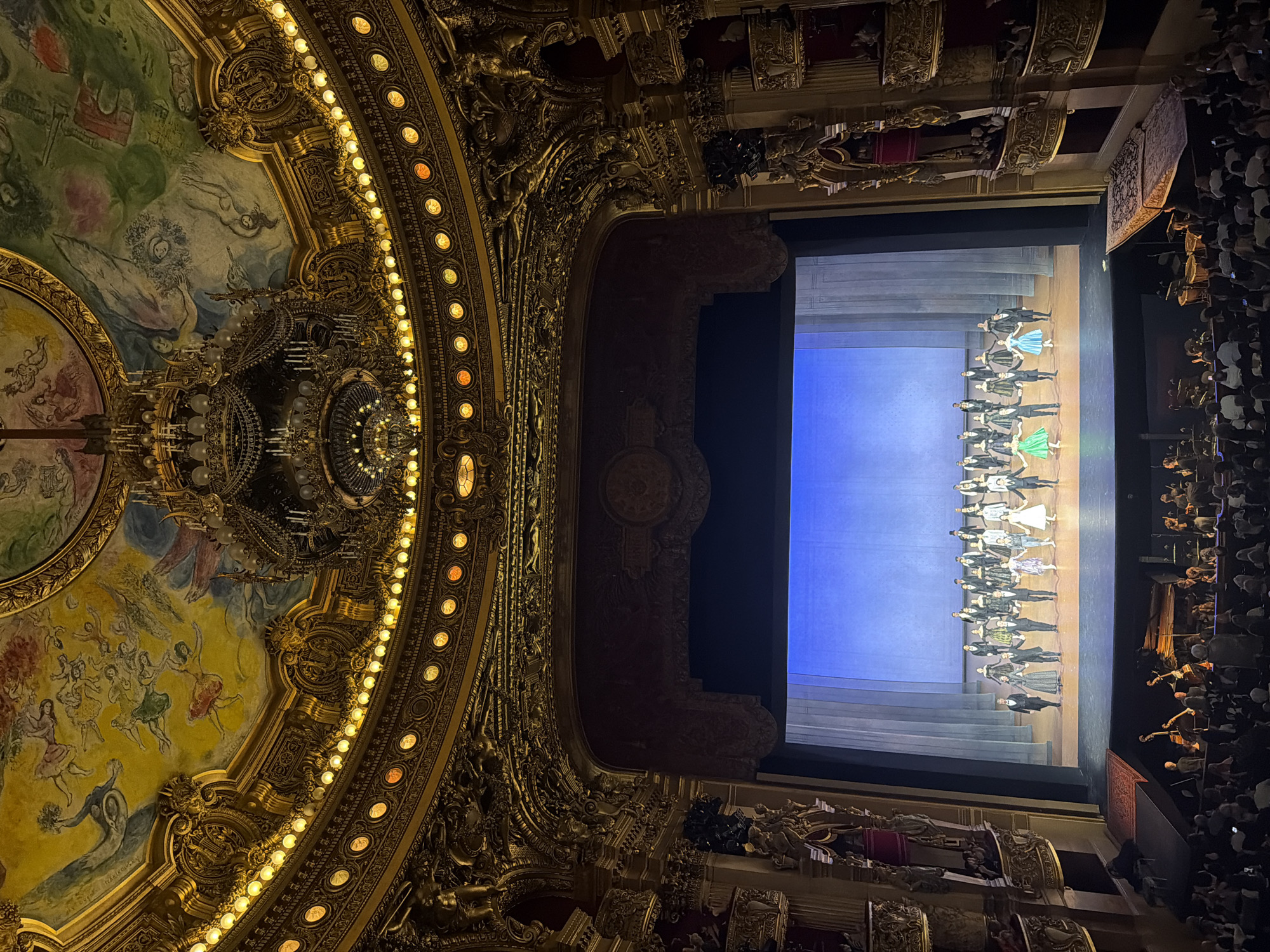 The image shows the inside of the Opéra Garnier in Paris, France, likely during a performance. The camera is pointed towards the stage, capturing the audience and performers. The stage is lit with a backdrop, and the performers are dressed in ballet attire, likely taking a bow. The interior of the opera house is ornate, with gold detailing, chandeliers, and a painted ceiling. The audience is visible in the foreground, filling the seats and balconies.