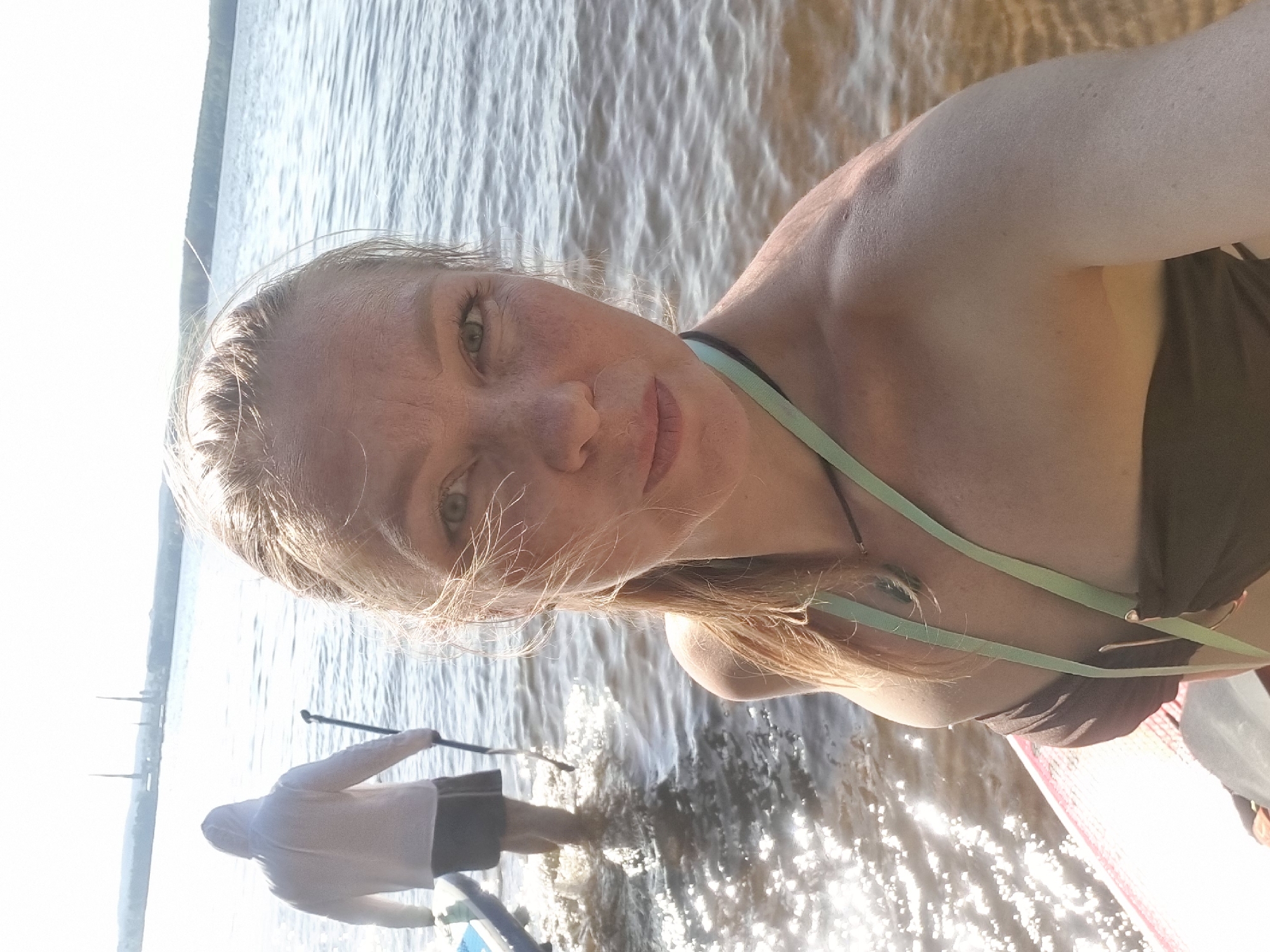 A woman is taking a selfie while stand-up paddleboarding on the water. Another person can be seen in the background, also on a stand-up paddleboard. The water appears to be a lake or river, with reflections of sunlight on its surface. The image was taken in the city of Chaykovsky, Russia.