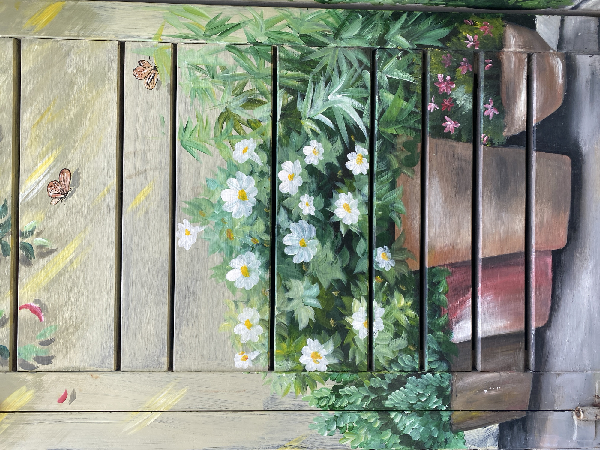 The image displays a close-up view of a painted shutter, likely on a building, featuring a garden scene. The shutter is composed of horizontal wooden slats. The painting depicts various elements of a garden, including green foliage, white flowers with yellow centers, and two small orange butterflies. In the foreground, there are several painted terracotta pots containing plants and flowers. The overall style is illustrative and somewhat impressionistic. The image does not provide any specific visual cues that would allow for identification of a location within Đà Nẵng, Vietnam.
