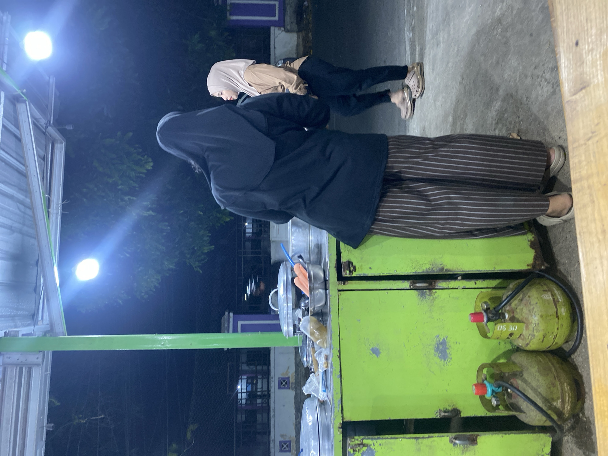 The image depicts an outdoor scene at night in Banjarbaru, Indonesia, likely near a street food vendor. In the foreground, two green LPG gas cylinders with red and blue valves are visible at the base of a vibrant lime-green food cart. The cart is laden with various items, including large metal pots with lids, bowls containing food, and what appear to be vegetables. A person wearing a black hijab and striped pants stands with their back to the camera, facing the cart. To the right, another person in a light brown hijab and black pants is partially visible, looking towards the first person. The overall lighting suggests artificial illumination from a bright overhead light source, casting a bluish glow. The background shows a dark, blurry environment with hints of trees and structures, consistent with an urban or suburban setting at night.