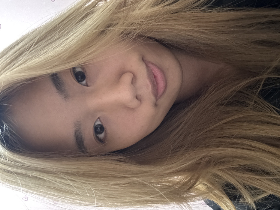 A close-up portrait depicts a young East Asian female in Shapingba District, China. The subject has long, light blonde hair, which frames her face and extends beyond the lower frame. Her skin tone is fair, and her dark eyes are open, gazing directly into the camera. Her lips are closed, forming a subtle, neutral expression. She is wearing a dark-colored garment, visible across her shoulders and upper chest. The background is a light-colored wall, possibly pinkish or off-white, adorned with faint, scattered small pink heart-shaped graphics, particularly visible in the upper left quadrant. The image is a selfie, taken from a slightly upward angle.