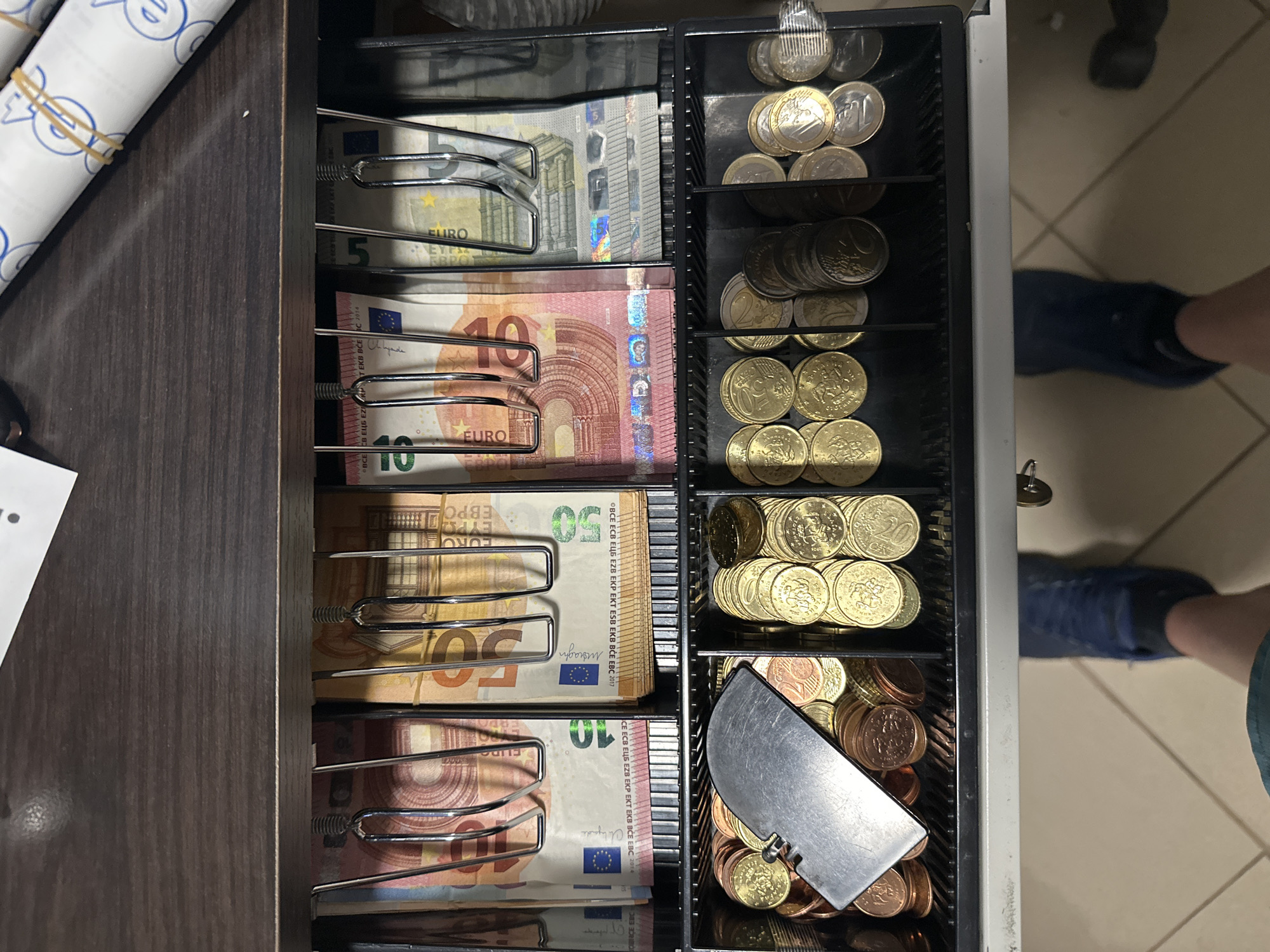 An overhead, rotated view captures an open cash drawer filled with Euro banknotes and coins, set within a dark wooden counter or cash register. To the left, several rolls of white paper with blue patterns, secured by elastic bands, rest on the wooden surface.

Inside the cash drawer, compartments are organized with Euro banknotes held by metal clips. Bundles of 5, 10, and 50 Euro notes are visible. Prominent texts on the banknotes include "EURO", "EBPO", and the denominations "5", "10", "50". Further details like "BCE ECB EZB EKP EKT EKB BCE EBC 2014" and "BCE ECB EZB EKP EKT EKB BCE EBC 2017" indicating the issuing central banks and year of series are also visible.

Adjacent compartments contain sorted Euro coins, including stacks of 2 Euro, 1 Euro, 50 cent, and 20 cent coins. The lowest compartment holds smaller copper coins, with a metal coin scoop resting among them.

The floor visible below and to the right of the drawer is tiled and light-colored. A person's feet, clad in dark blue shoes, are seen on the tiles, indicating someone is standing near the open till. The scene depicts a cash management activity, likely a transaction, in a commercial establishment in Ivailo, Bulgaria, where Euro currency is being used.