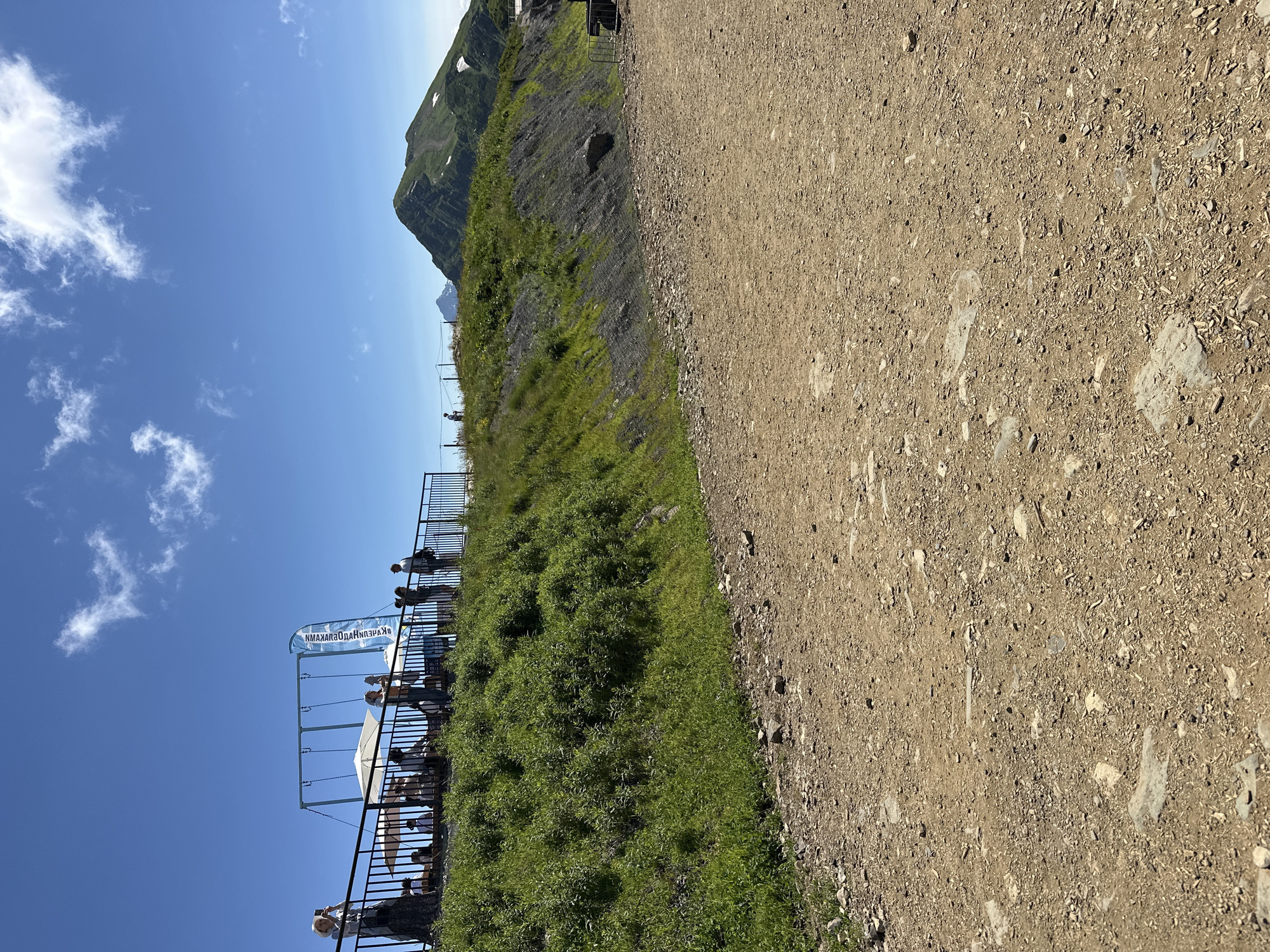 The image shows a view of a mountain landscape with a dirt path in the foreground and a grassy slope leading up to a viewing area with people. A distant mountain peak is visible in the background. The viewing area has a black metal fence, several people standing and sitting, some umbrellas, and a swing set. A blue banner with text is also present. The sky is clear blue with some scattered white clouds. The location is identified as being in Estosadok, Russia.