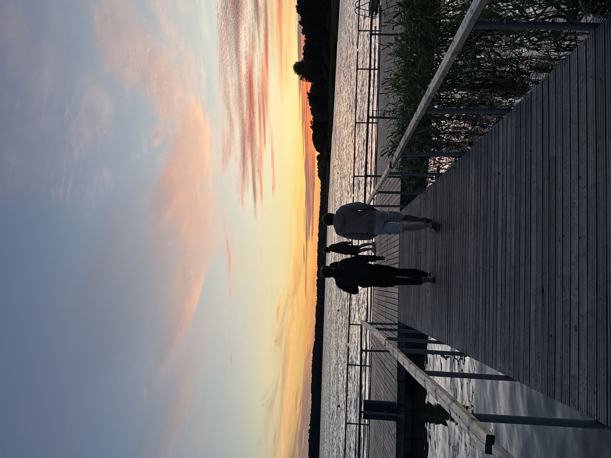 The image depicts a wooden pier extending into a body of water during sunset. The sky is painted with hues of orange, pink, and blue, indicating the sun is low on the horizon. The water is calm, reflecting the colors of the sky. On the pier, three individuals are walking away from the camera, their silhouettes visible against the vibrant sky. Two of the individuals are walking side-by-side, while the third is slightly behind and to the left. The pier has wooden planks and metal railings. On the right side of the pier, tall grass or reeds are growing near the water's edge. The background shows a treeline on the far side of the water. There is no identifiable indication that this scene is located in Tallinn, Estonia.