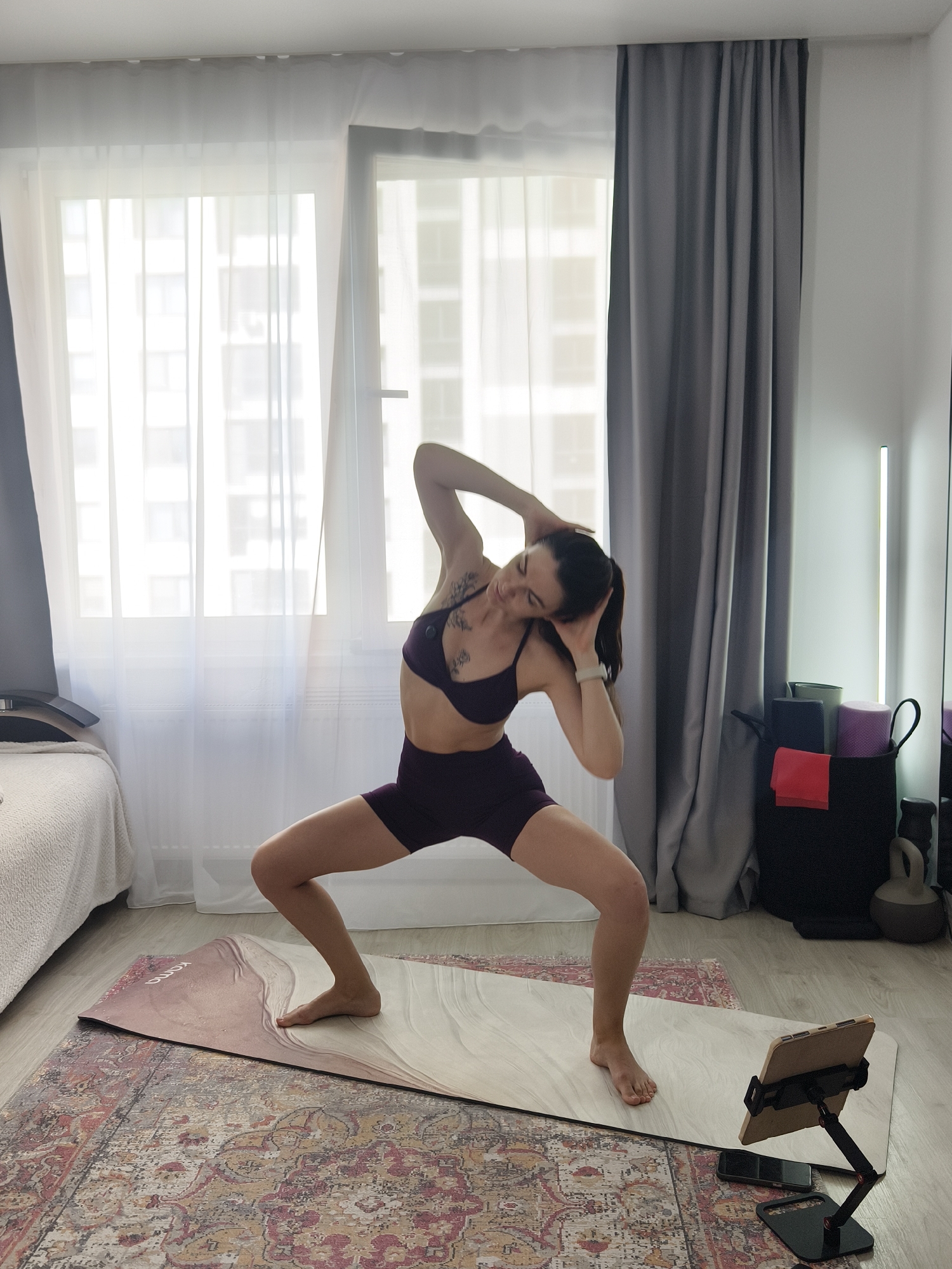 A young woman is performing a yoga pose on a mat in a well-lit room. She is wearing a dark purple sports bra and shorts, and her hair is in a ponytail. Her right hand is behind her head, and her left arm is extended to the side. Her legs are bent, and her feet are flat on the mat. The room has large windows with sheer curtains, and a gray curtain hangs on the right side. There is a bed on the left side of the room, and a storage bin filled with yoga equipment, including blocks and rolled-up mats, is on the right. A tablet on a stand and a smartphone are placed on the floor in front of the woman. The overall impression is of a home yoga practice.
The location of this image is not specified.