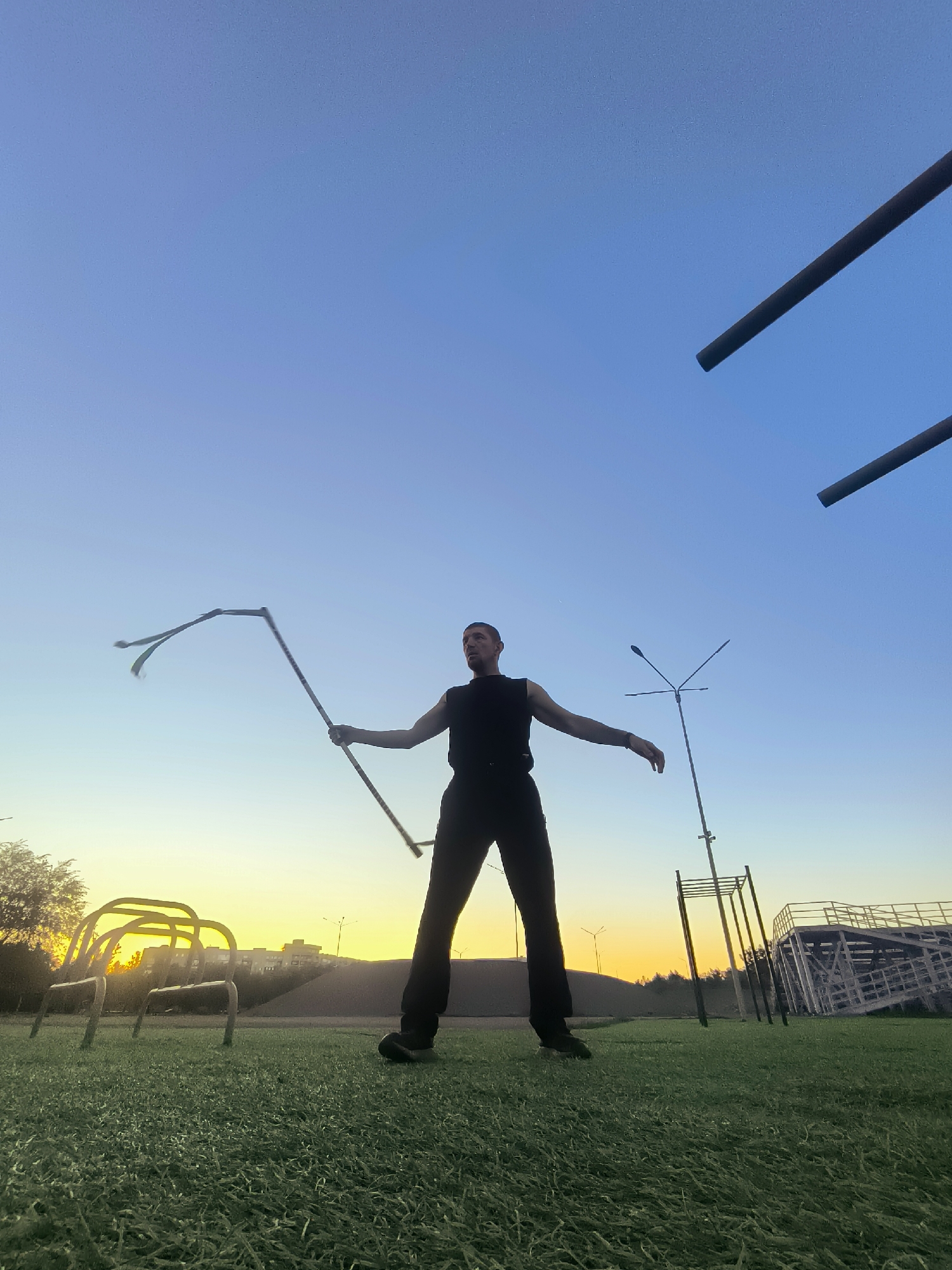A man is shown practicing with a prop that has streamers attached to it. The man is wearing a black sleeveless shirt and black pants. He is standing in a grassy field with a clear blue sky above him. In the background, there are some playground equipment, a lamp post, and buildings. The sun is setting, casting a warm glow over the scene. The location is the city of Qarağandı, Kazakhstan.