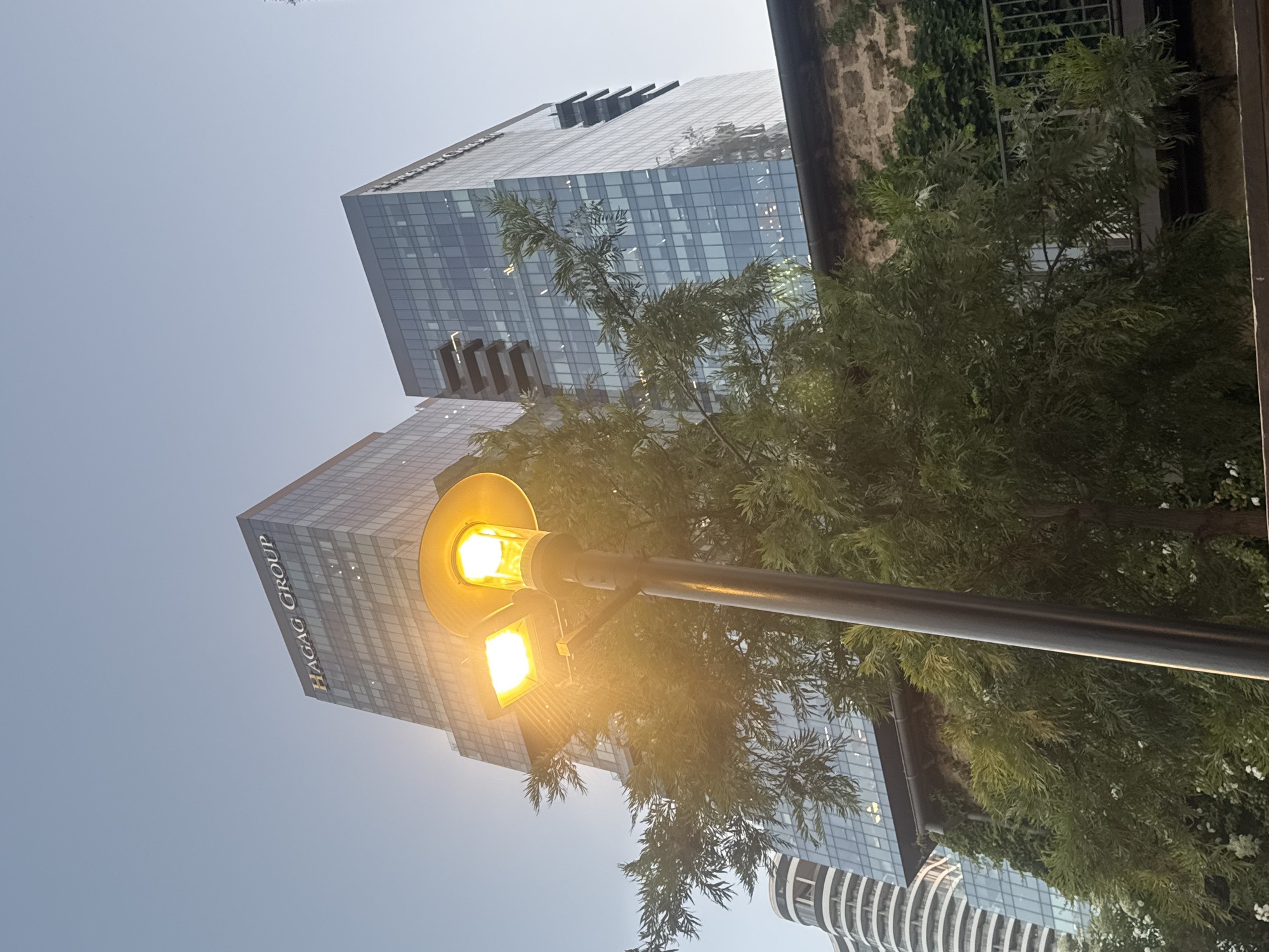 The image presents a tilted, upward view of a modern urban landscape in Tel-Aviv, Israel, under a bright, clear blue sky.
The primary subjects are two contemporary glass skyscrapers, a lamppost with illuminated lights, and lush green foliage. The building on the left features the prominent, vertically oriented text "HAGAG GROUP" on its side, indicative of a corporate structure. Both skyscrapers are characterized by their reflective glass facades and grid-like window patterns, with the one on the right displaying dark, rectangular architectural elements protruding from its upper sections.
A black lamppost stands prominently in the foreground, equipped with two bright, warm-toned lights—one circular and one rectangular—that are illuminated despite the apparent daylight, creating a strong glow and lens flare effect. Various green trees and bushes partially obstruct the view, their leaves appearing vibrant and healthy. In the far right background, a contrasting older structure with rough, stone-like walls and climbing ivy is visible, adjacent to a dark, possibly metal, grate. A section of a third, distinct modern building with curved, striped details can be seen at the bottom right.
No specific activity or people are depicted; the scene is static, focusing on architecture and nature. The clear sky and bright illumination suggest a sunny day, likely during daylight hours.