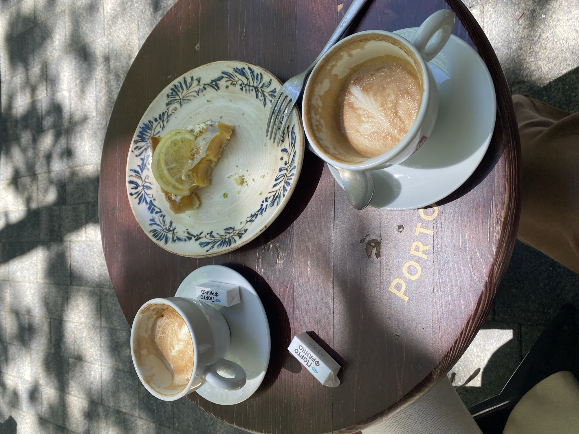 An overhead photograph captures a sun-dappled outdoor café table in Sochi, Russia. The round, dark wooden table features a partially consumed snack or meal. On a white plate with blue, floral-like patterns, a slice of lemon tart, topped with a lemon wedge and cream, is visible with a silver fork resting on it.
Two white coffee cups, each with a matching saucer, are also on the table. Both cups contain remnants of coffee, with faint latte art designs still discernible in the foam. Two white sugar packets are placed near the lower coffee cup. Each packet prominently displays "ПОРТО ФРАНКО" (Porto Franco) in blue Cyrillic script, accompanied by a small boat logo.
The table itself has "PORTO" inscribed in gold lettering along its right edge. The surrounding ground, visible beyond the table, consists of grey paving stones, indicating an outdoor setting. Strong, dappled sunlight casts distinct shadows of leaves or branches onto both the paving stones and parts of the table, suggesting a bright and sunny day. The scene conveys a moment of relaxation during a coffee and dessert break.