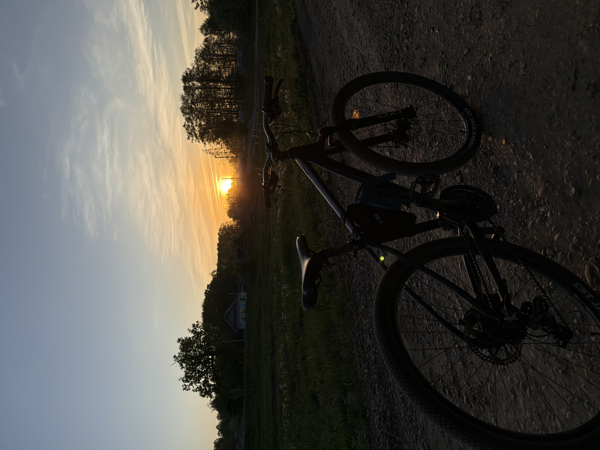 A bicycle is positioned on a dirt road during sunset. The sun is low on the horizon, obscured by trees and clouds. The surrounding environment includes trees and a building, consistent with the location of Noviy, Russia.