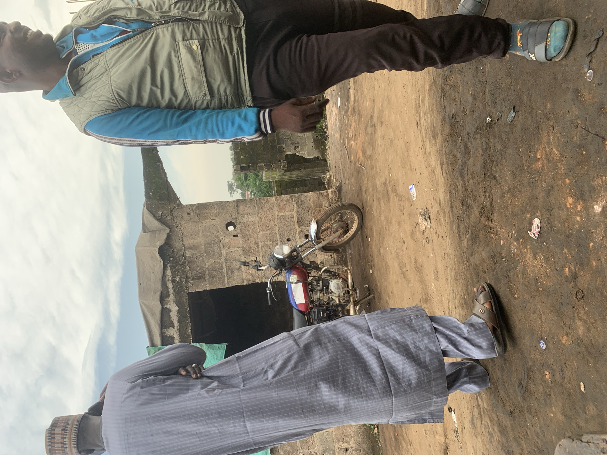The image features two individuals in an outdoor setting, likely a somewhat dilapidated urban or semi-urban area in Shagamu, Nigeria. The foreground is dominated by the lower half of a person wearing a grey pinstriped garment, a cap, and sandals. To the left and slightly behind this person, another individual is visible from the waist up, wearing a green puffer vest over a blue long-sleeved shirt and dark pants. This second person is smiling and appears to be looking upwards.
Between the two individuals, a motorcycle is parked. It is red and blue with a silver engine and a visible license plate. The background consists of rough brick structures, one of which has a dark opening. The ground is dirt and appears somewhat muddy and littered with debris, including wrappers and small discarded items. The sky is overcast with a pale, diffused light, suggesting it is daytime, possibly morning or afternoon, with no direct sunlight. The overall impression is that of an everyday scene in a non-pristine environment. No text is clearly discernible in the image.