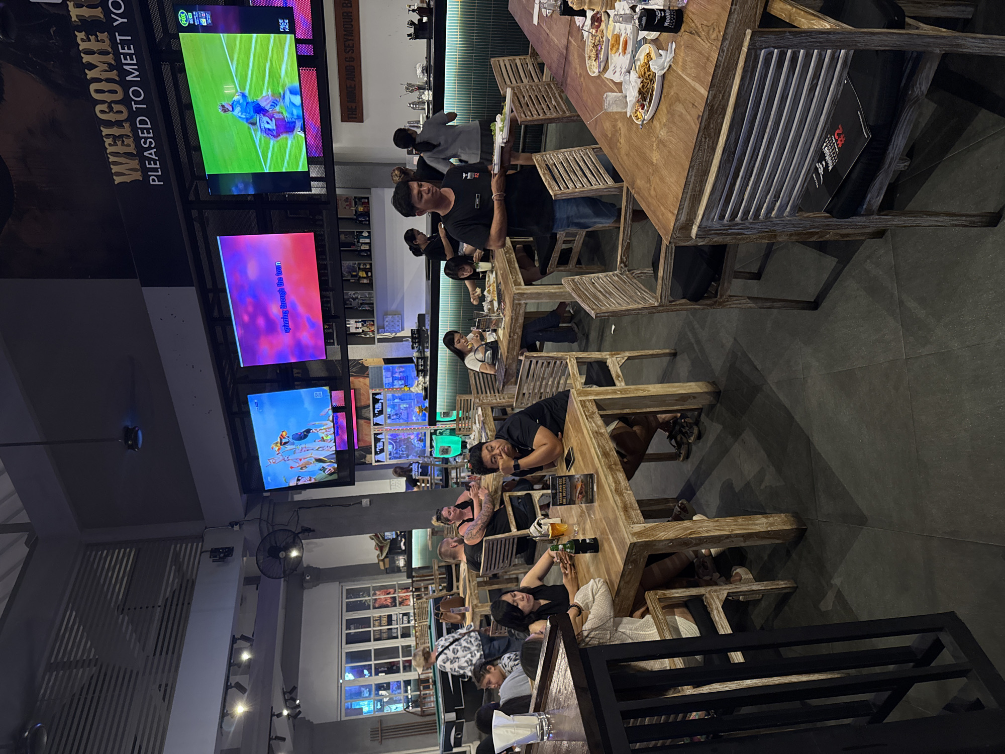 The image shows the interior of a busy restaurant or bar in Kuta, Indonesia. Multiple large screens are mounted on the wall, displaying sports or entertainment. Several tables with diners are visible, with people eating, drinking, and socializing. A server is carrying a tray of food. The decor includes wooden furniture and a bar area with bottles displayed. The atmosphere appears casual and lively.