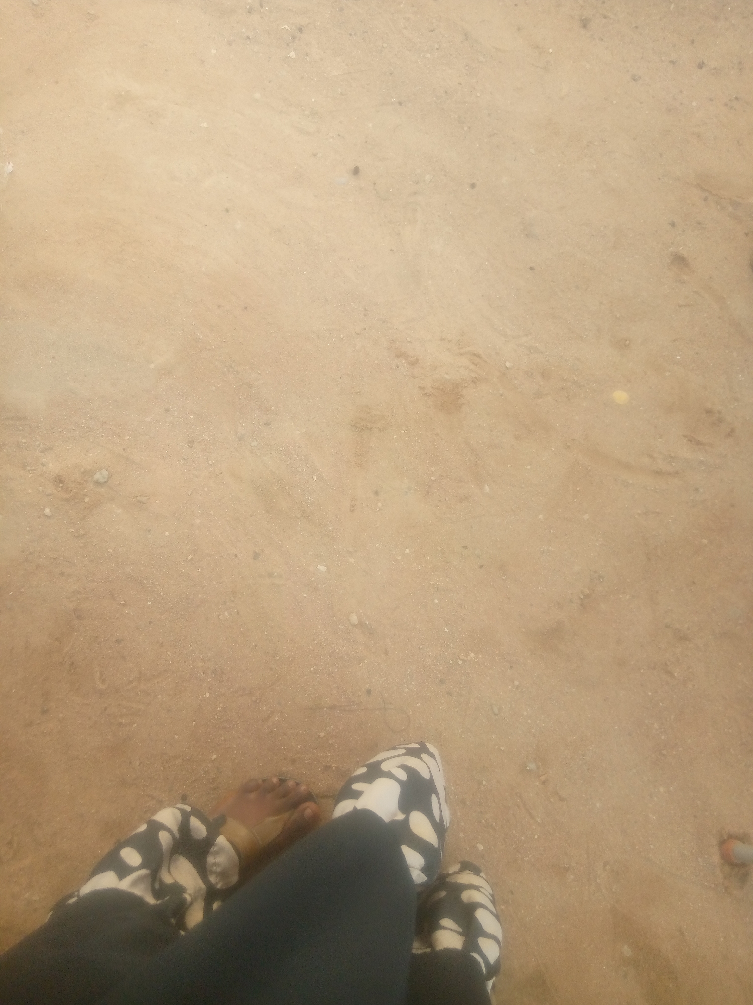 The image shows a close-up view of two feet standing on a sandy, dry ground. The feet are wearing patterned shoes with a black and white design. One foot is partially obscured by dark clothing. The ground appears to be a natural surface, possibly a dirt path or undeveloped area, with small pebbles and debris scattered across it. The lighting suggests an outdoor environment. The scene is devoid of any identifiable city landmarks or structures, making it impossible to ascertain a specific location within Jalingo, Nigeria.