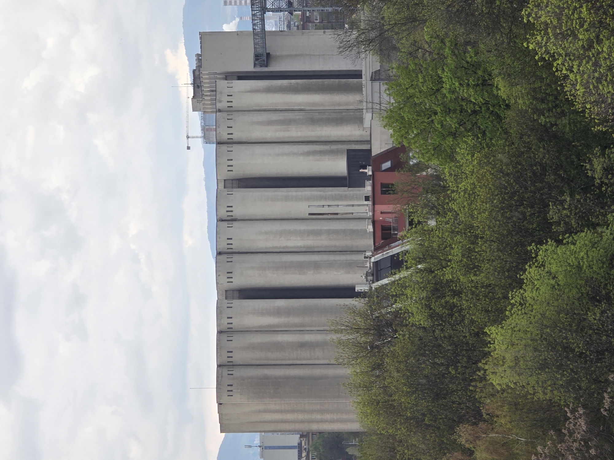 The image captures an industrial and natural landscape under a cloudy daytime sky, likely in Sofia, Bulgaria. The scene is dominated by a series of imposing, tall, grey concrete structures, resembling silos or part of a large industrial complex, positioned towards the right side of the frame. These structures feature numerous small, uniformly spaced rectangular openings or windows along their length, and display signs of weathering with darker vertical streaks.
In the foreground and partially obscuring the lower sections of the concrete buildings, there is a dense expanse of vibrant green trees, indicating spring or early summer foliage. Amidst the greenery and in front of the grey silos, a small, distinctive red-orange building with dark windows stands out.
Above and behind the main concrete structures, elements of further industrial infrastructure are visible, including parts of a large metal gantry or bridge-like structure and a construction crane, hinting at ongoing development or operations. The sky occupies a significant portion of the left side of the image, filled with soft white and light grey clouds, suggesting an overcast but bright day. Faint outlines of distant mountains or hills can be seen on the horizon.
No people or specific activities are visible, presenting a static view of the environment. There is no discernible text within the image. The overall impression is one of an industrial area situated alongside or within a verdant natural setting.