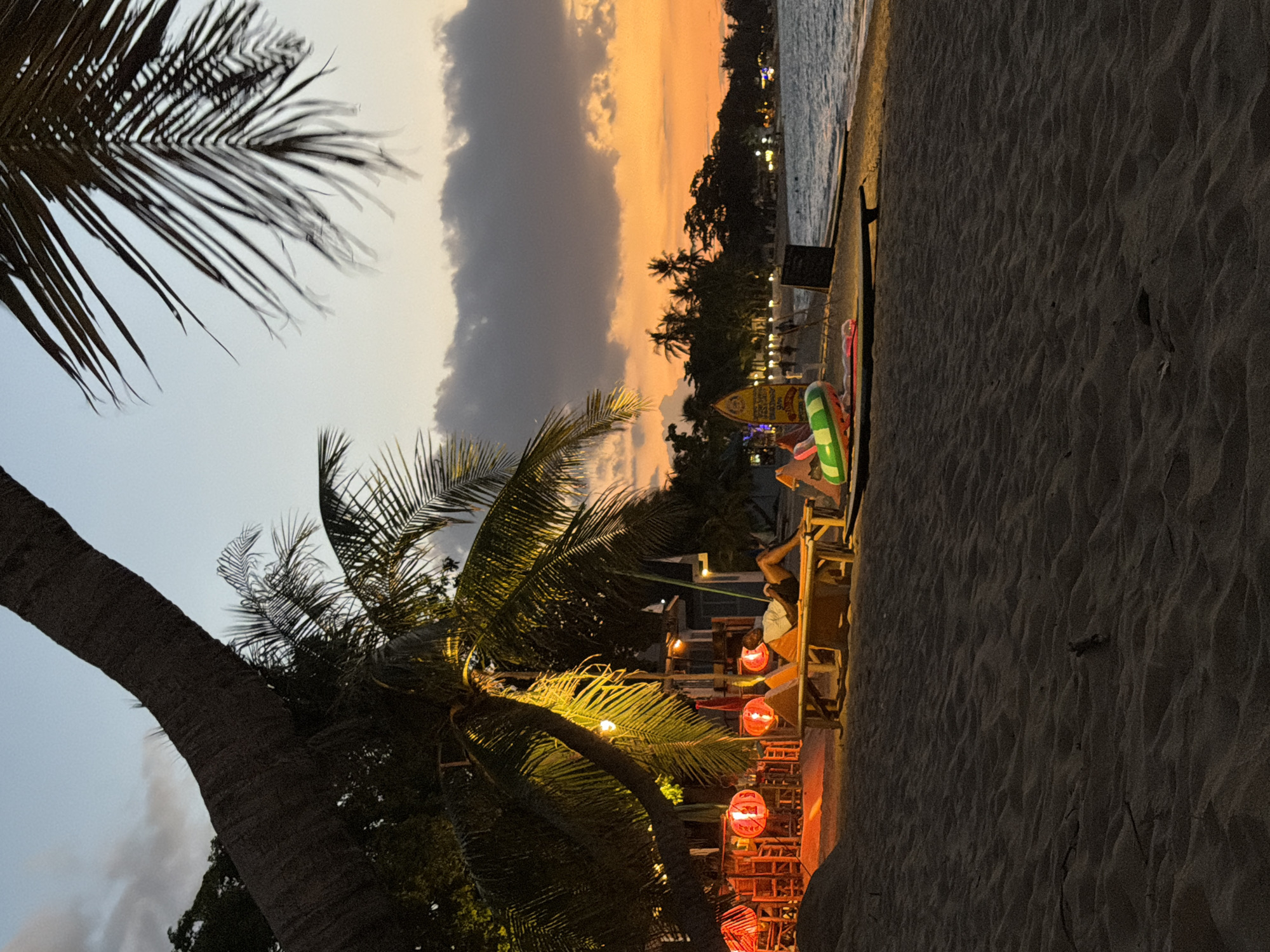 A beach scene at dusk in the Ko Samui District of Thailand. A large palm tree dominates the left foreground, its trunk curving upwards and its fronds casting shadows. Underneath the tree, a beachside restaurant is illuminated by red lanterns, with tables and chairs visible. In the mid-ground, two individuals are relaxing on lounge chairs on the sand. To the right, the ocean stretches out, with gentle waves lapping the shore. Palm trees and buildings line the distant coastline, lit by artificial lights. The sky is a gradient of orange, yellow, and grey clouds, indicating sunset. Beach toys, including an inflatable ring, are scattered on the sand.