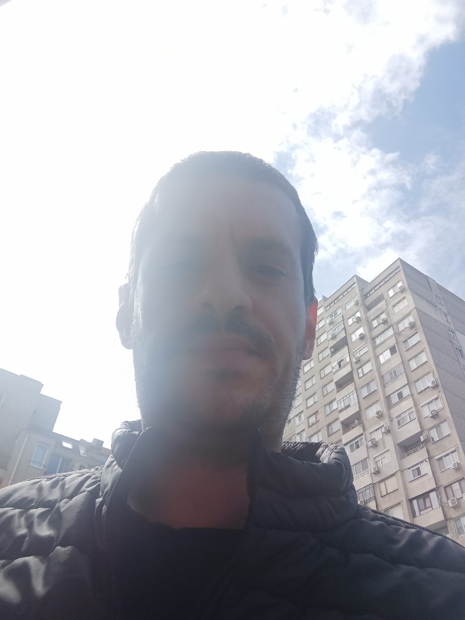 A selfie-style, upward-angled photograph shows a man with dark hair and a mustache, wearing a dark jacket. The background includes a cloudy sky and an older apartment building with numerous windows and air conditioning units. This building is located in Burgas, Bulgaria. Other buildings can be seen in the distance.