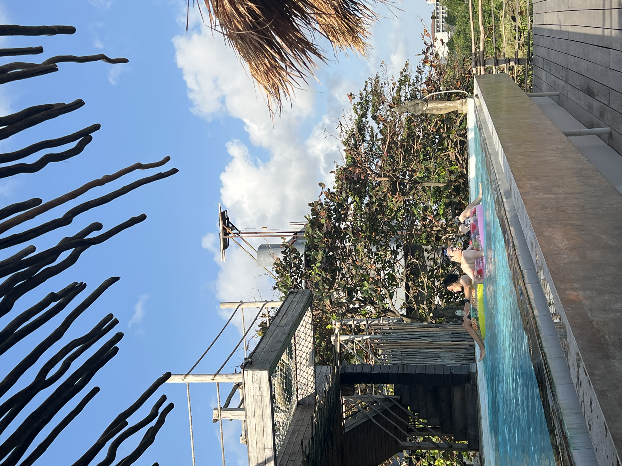 The image depicts an outdoor infinity pool in Mengwi, Indonesia, likely at a resort or private residence, with two individuals relaxing in the water. The pool is situated on an elevated platform, with wooden decking visible in the foreground. Palm fronds overhang from the top of the frame, creating a natural canopy. Behind the pool, lush green trees and vegetation are abundant, with buildings visible in the distant background. A statue stands near the edge of the pool. The sky is a vibrant blue with scattered white clouds. The two people are each in an inflatable tube, one in a yellow tube and the other in a pink tube, both appearing to be casually enjoying the water. The overall atmosphere suggests a tranquil and tropical setting.