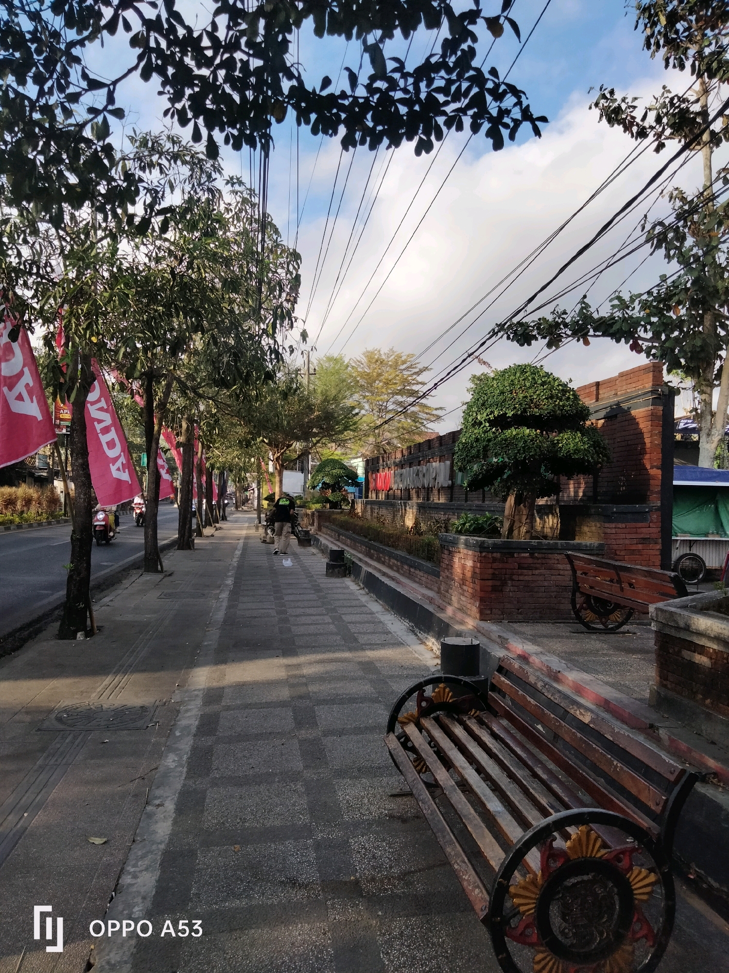 A street scene in Kelutan, Indonesia, shows a sidewalk lined with trees bearing red flags. Several people are walking on the sidewalk. One person is pushing a stroller. In the background, there is a brick wall with the words "Taman Wisata" written on it. There are also several benches on the sidewalk. The sun is shining, and the sky is blue with some clouds. The scene appears to be peaceful and serene.
