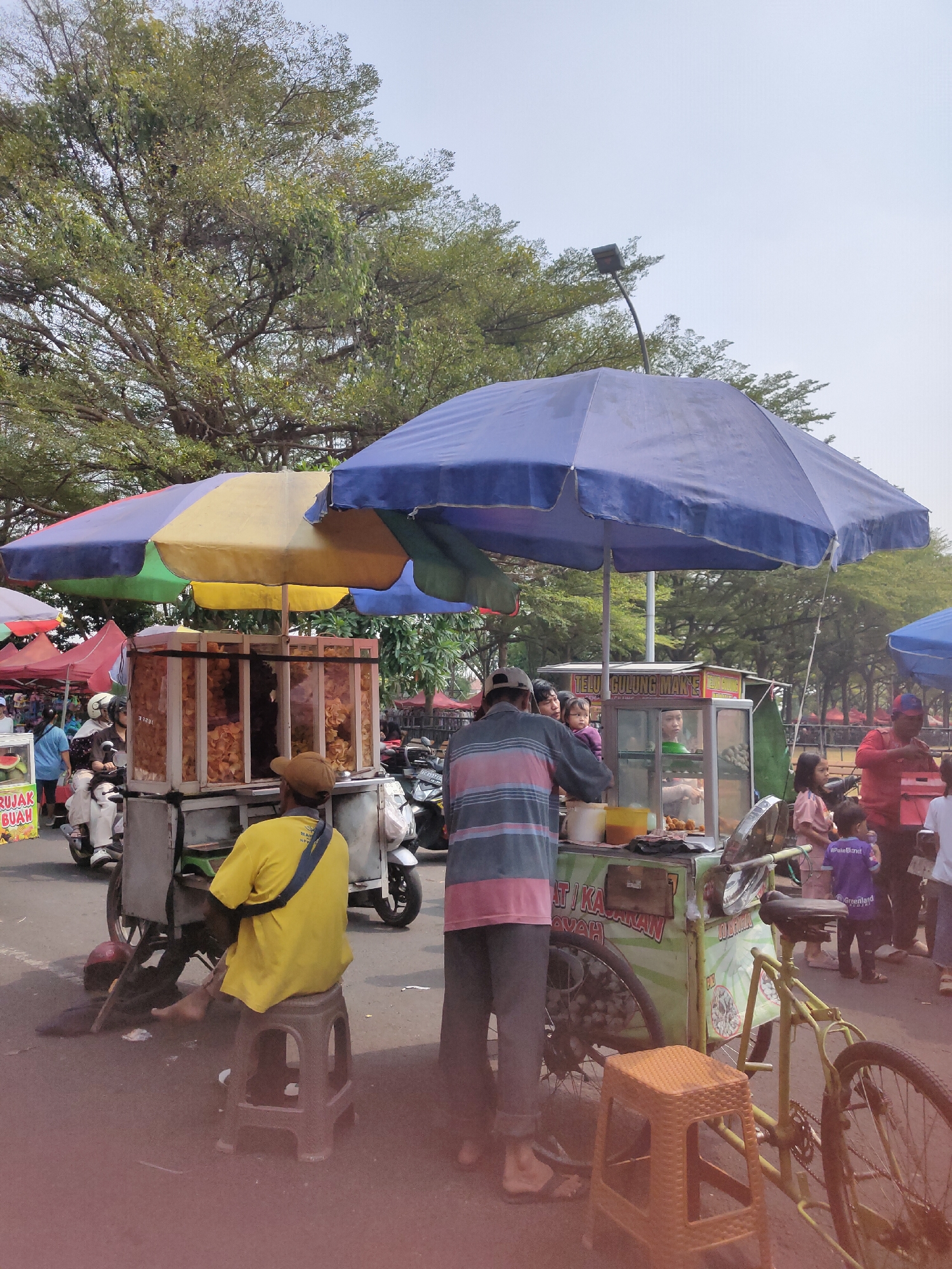 In Ngasem, Indonesia, an outdoor market scene unfolds under a bright sky, with lush green trees forming a canopy above. A variety of food stalls line the paved ground, adorned with colorful umbrellas. In the foreground, a man in a yellow t-shirt and brown hat sits on a stool, facing away from the camera. He appears to be tending to a food cart, possibly a mobile kitchen on wheels.
To his right, another food cart is visible, featuring a glass display case filled with snacks and items for sale. A man in a striped shirt stands by this cart, facing the viewer. Other people are scattered throughout the scene, including a woman holding a child, and several children in the background. Motorcycles and bicycles are parked nearby, suggesting a busy marketplace. The overall atmosphere appears lively and active, typical of a bustling Indonesian street market.