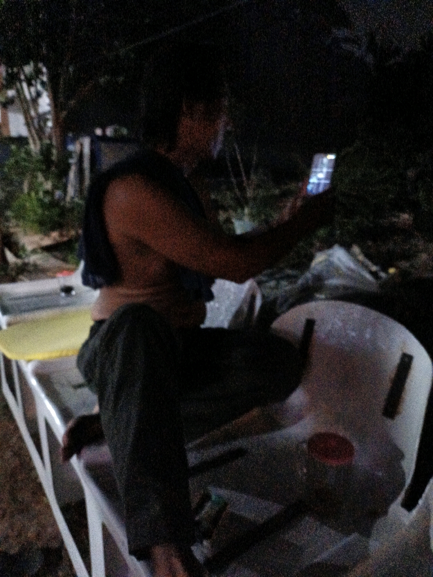 The image depicts a dimly lit outdoor scene, likely at night, given the low light conditions and the glowing screen of a smartphone held by a person in the foreground. The person, whose body is partially visible from the side and back, appears to be seated and engaged with the phone. They are wearing dark-colored clothing, possibly pants and a t-shirt or tank top, with a dark fabric draped over their shoulders. The background suggests an area with some vegetation, trees, and possibly some structures or discarded materials, contributing to a somewhat cluttered environment. The image quality is grainy, which is characteristic of low-light photography. The presence of a white plastic chair, a yellow surface, and some white and translucent plastic objects in the foreground indicates a potentially public or communal outdoor space, such as a park, roadside rest area, or a gathering spot. Without specific landmarks or contextual clues within the image, pinpointing the exact location in Cikal, Indonesia, is not possible. However, the general setting suggests an urban or semi-urban environment in Indonesia, characterized by typical outdoor furniture and possibly a less-than-pristine surrounding.