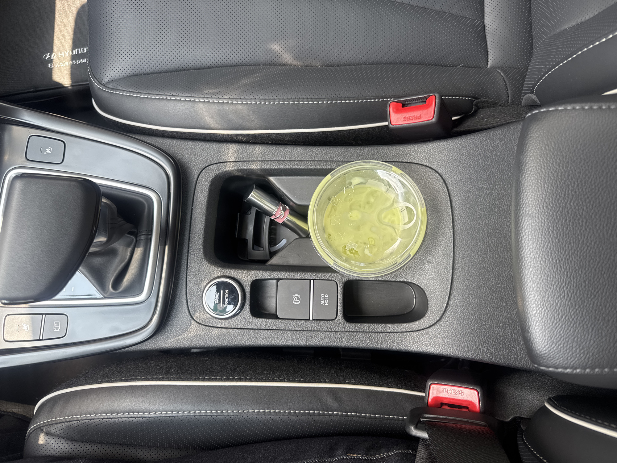 A top-down shot shows the interior of a car. The center console between the two front seats holds a clear plastic cup filled with a green liquid. Beside the cup is a slim, possibly metallic, object with a pink accent. Below these items are car controls: "DRIVE TRACTION", an electronic parking brake button marked with a "P", and an "AUTO HOLD" button. The black leather seats are visible on either side, marked with white stitching along the edges. The seatbelts are buckled, showing red "PRESS" buttons on each side. On the left, there is a portion of the gear shift. A car floor mat with the Hyundai logo is partially visible near the left seat. The specific location in the city of Puebla, Mexico, is not discernible from the image.