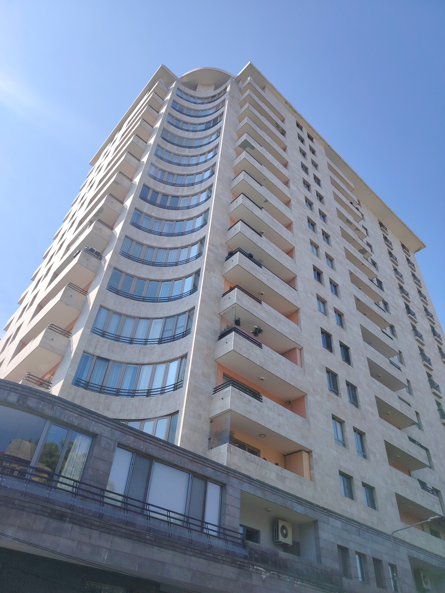 This is a low-angle, full-frame photograph of a modern apartment building in Yerevan, Armenia. The building is tall and has a light beige facade, likely made of stone tiles. It features a curved section on the left side with a series of large, arched windows that appear to be tinted. The right side of the building has a more rectilinear design with numerous rectangular windows and balconies. The balconies are mostly open with dark railings and some have planters. The lower portion of the building is clad in a darker, grey stone. The sky is a clear, bright blue with no clouds. The sun is out, casting shadows and creating a lens flare in the upper left corner. The image is taken from ground level looking upwards at the structure. There are no people visible in the image. The overall impression is of a contemporary urban residential building.