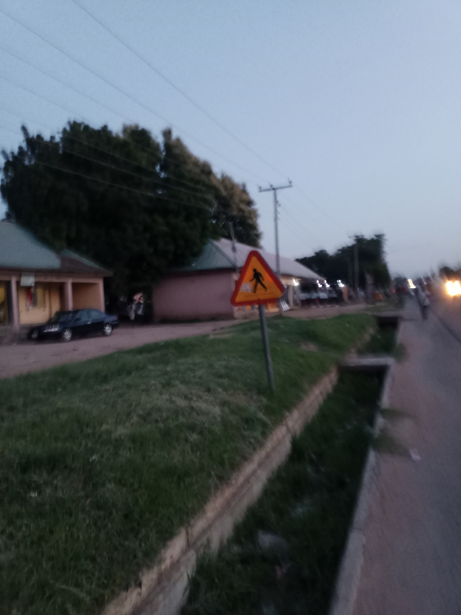 The image depicts a street scene in Jalingo, Nigeria, likely at dusk or dawn due to the low light conditions. In the foreground, there is a grassy area with a drainage ditch running alongside a paved road. A yellow triangular warning sign with a silhouette of a person walking is visible on a pole.
On the left side of the road, there are buildings with a dark-colored sedan parked in front of one. The buildings appear to be single-story structures. Further down the road, more buildings and streetlights can be seen. Several people are walking on the road, and a vehicle with illuminated headlights is present in the distance. The environment suggests an urban or semi-urban setting with residential or commercial structures lining the street. Power lines are visible stretching across the sky above the trees.