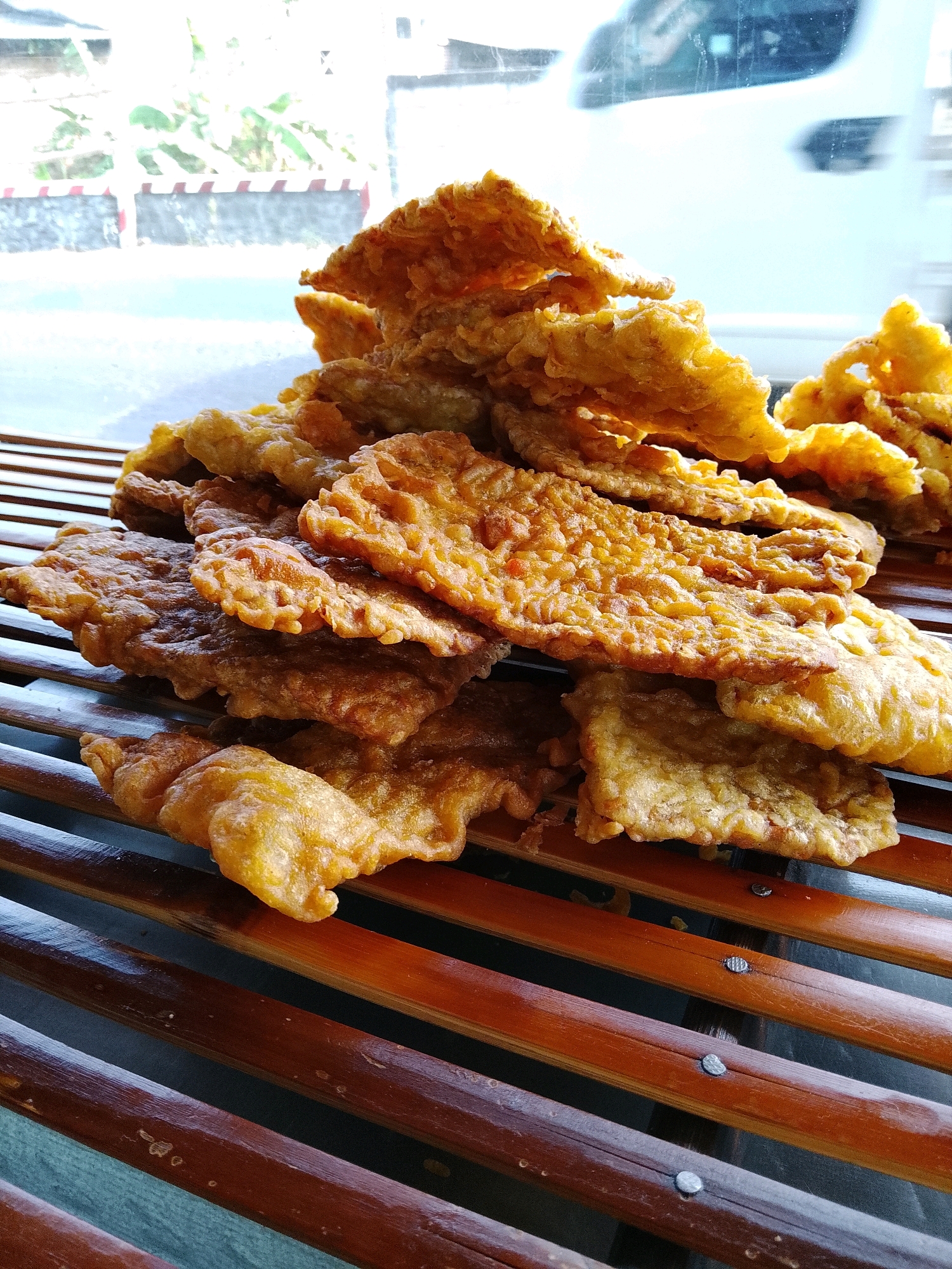 The image shows a close-up, slightly angled shot of a pile of golden-brown fried food, likely tempeh goreng, arranged on a slatted wooden surface. The fried pieces are irregularly shaped, with a crispy, textured coating. In the background, through a window, a blurred white vehicle is visible, suggesting it is on a street. To the left, a red and white striped barrier and some greenery are also faintly discernible. The wooden slats appear to be a light to medium brown color, with some variation in tone and texture. The lighting suggests an outdoor or well-lit indoor setting, casting subtle shadows on the food. The overall impression is one of food being displayed or prepared for sale, possibly in a casual setting. The image is identified as being taken in Banaran, Indonesia.