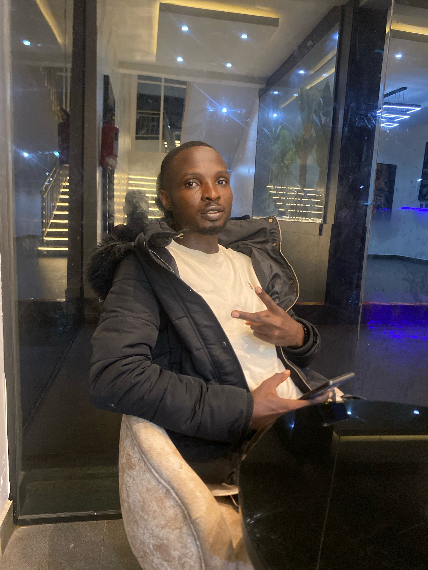 A man in a black puffer jacket with a fur-lined hood and a white t-shirt sits in a chair at a dark, reflective table. He is looking towards the camera and holding his left hand up with two fingers extended in a peace sign. His right hand is holding a smartphone. The background appears to be an indoor setting with glass walls reflecting stairs with lights and some greenery. The location is in the city of Barden Yana, Nigeria.
