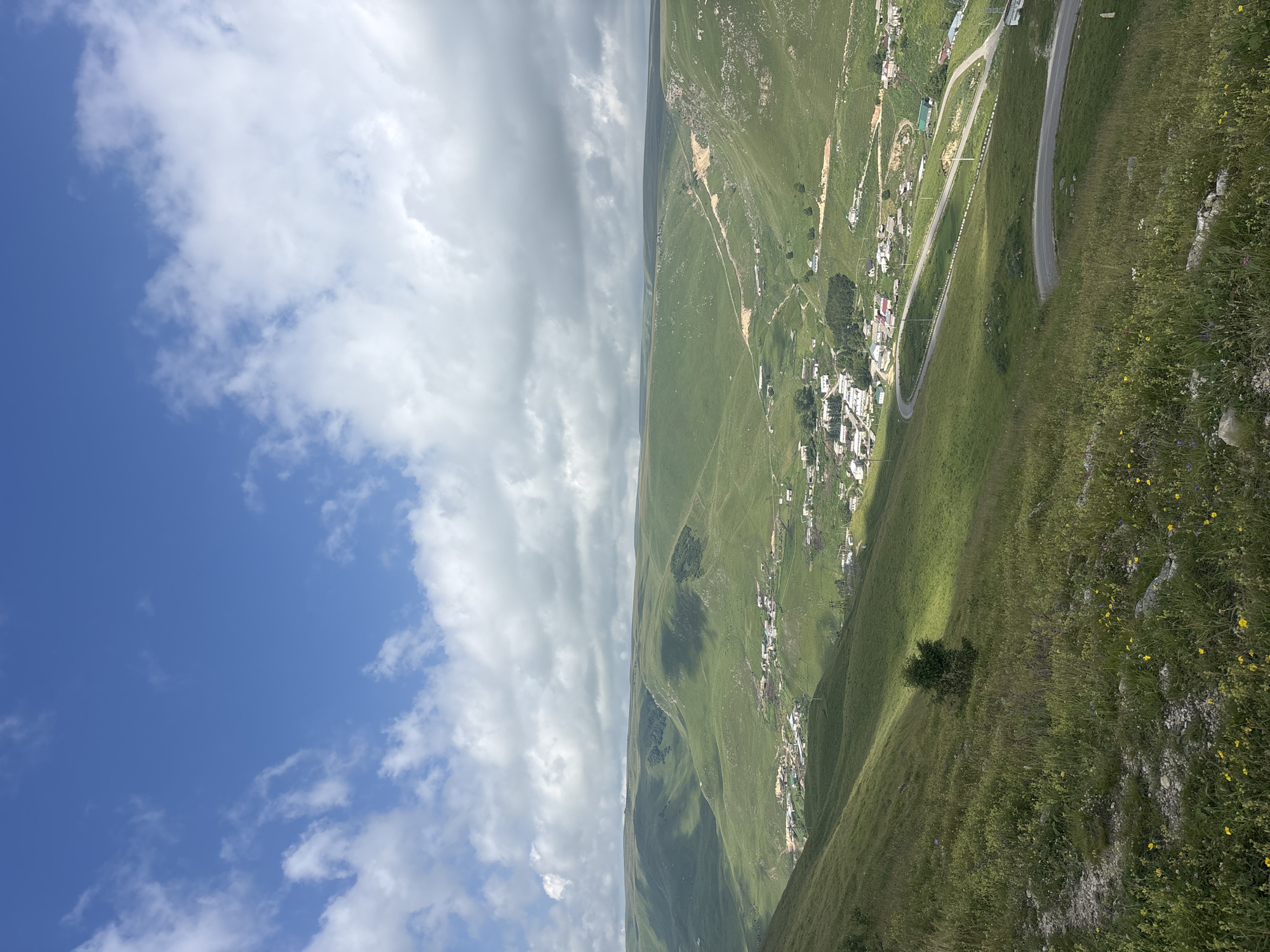 The image captures a vast, rolling green landscape under a bright blue sky with scattered white clouds. In the mid-ground, a small village is nestled in a valley, with buildings clustered together and a winding road passing through it. The road is paved and has white lines marking the lanes. Power lines and poles are visible along the road and in the distance. The foreground consists of a grassy slope dotted with small yellow wildflowers and some rocks. The overall scene depicts a rural, mountainous environment in the vicinity of Kichi-Balyk, Russia. There are no people visible in the image. The lighting suggests it is daytime, likely mid-morning or afternoon, with shadows indicating the sun is at an angle.