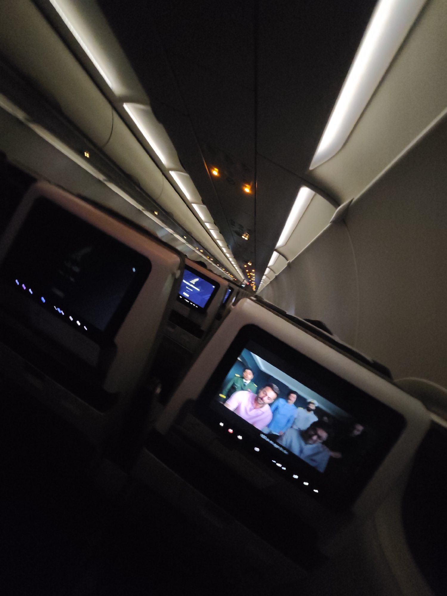 The image depicts the interior of an airplane cabin, likely during a flight. The perspective is from a passenger's seat, looking forward and slightly upwards towards the ceiling. The cabin is dimly lit, with the primary illumination coming from the overhead light panels that run along the length of the fuselage.
In the foreground, a personal in-flight entertainment screen is visible, displaying a scene from a movie or show featuring several men. The screen is angled towards the viewer. To the left, another, larger screen is visible, also dark, suggesting it is either off or displaying a menu. Rows of small, colored lights are also present below the left screen.
The ceiling of the cabin features a series of recessed light fixtures, with some illuminated in a cool white light and others appearing darker. The overhead compartments are not clearly visible but their presence is implied by the curvature of the cabin's interior.
The overall environment suggests a typical commercial airplane cabin during a flight, potentially at night or in a darkened cabin setting. There is no indication of the city of Puttaparthi, India, or any specific location within a city in the image.