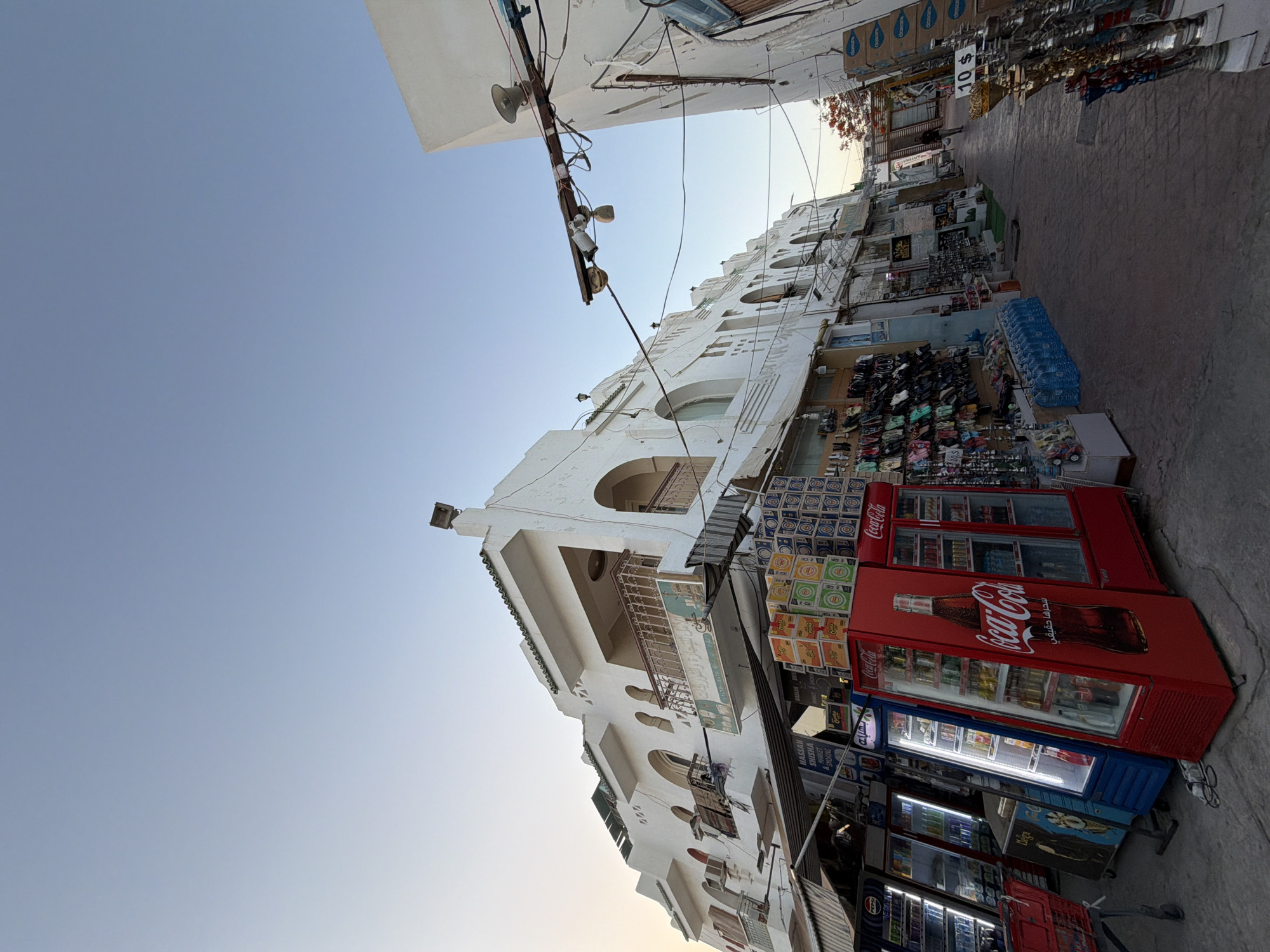 This is a vertical, low-angle shot of a bustling street scene in Sharm El Sheikh, Egypt, during the daytime. The scene features a narrow street lined with white buildings on the left and shops on the right. The shops display a variety of goods, including shoes, clothing, and drinks, with several refrigerators prominently featuring Coca-Cola branding.
On the left, the white buildings have a distinctive architectural style with arched windows and balconies, some adorned with ornate railings. Overhead, electrical wires and street lights are visible, adding to the urban feel. The sky above is a clear, pale blue, suggesting fair weather.
On the right side of the street, various vendors are set up. In the foreground, there are several large, red Coca-Cola refrigerators, one displaying a prominent image of a Coca-Cola bottle and Arabic text. Adjacent to these are other refrigerators, some lit up, and shelves stacked with bottled water and other merchandise. Above the shops, signs and displays are visible, including one with "10 $" indicating pricing. Arabic text is present on some of the signs and product packaging.
The overall impression is of a lively marketplace, likely a tourist area, given the type of goods on display and the prominent international branding. The low angle makes the buildings appear tall and imposing, while the focus on the shops and their contents highlights the commercial activity of the location. The time of day appears to be late afternoon or early evening, judging by the soft lighting and the clear sky.
Visible text includes "Coca-Cola," "10 $," and Arabic lettering on various signs and products.