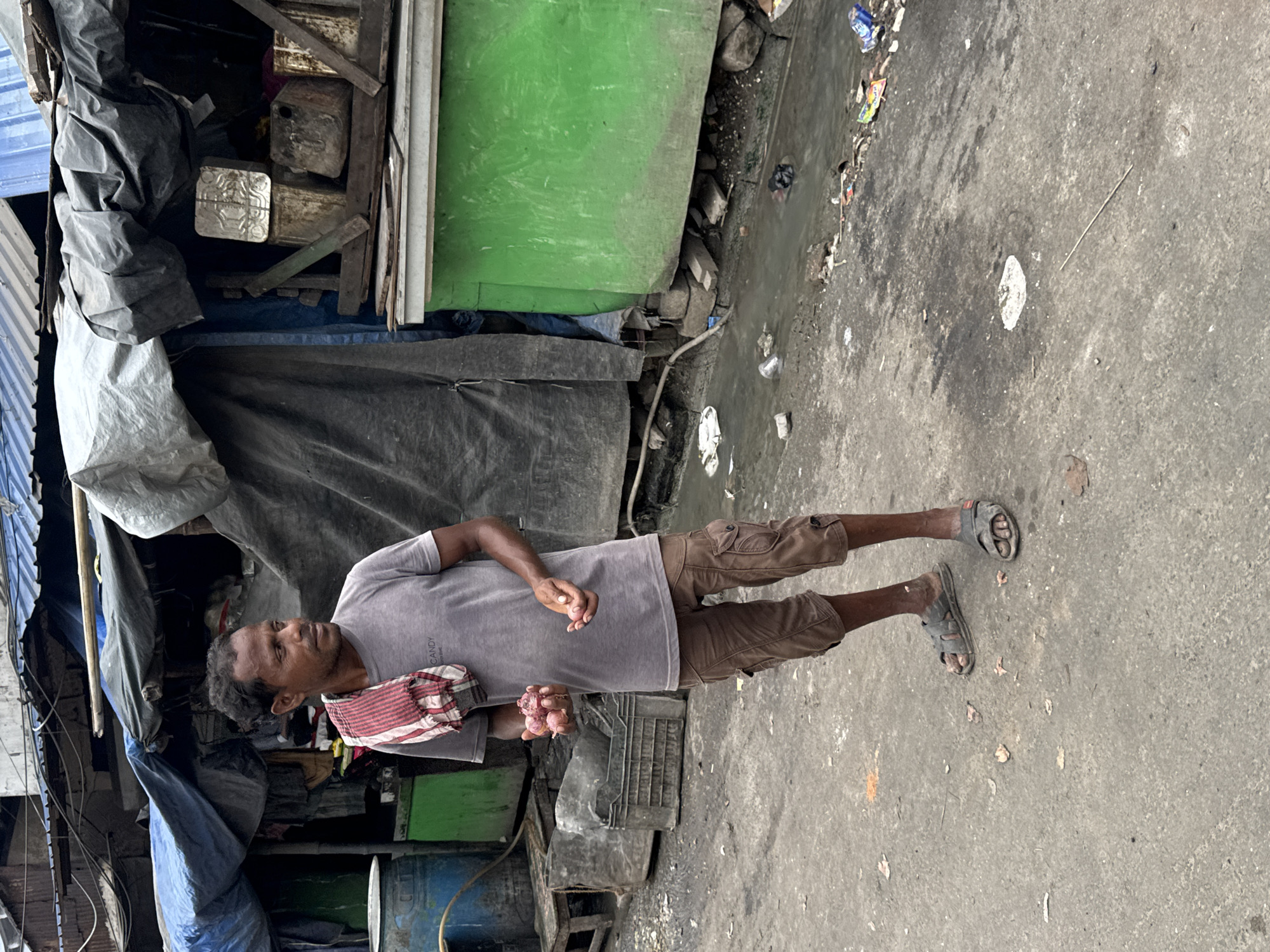 A man stands in a dirty alleyway, holding red onions in his left hand and a single onion in his right. He wears a grey t-shirt and brown cargo shorts, with a red and white striped cloth draped over his shoulders. The background features makeshift stalls with tarpaulin coverings, some with green painted fronts. A shallow, muddy trench with debris runs along the side of the alley, suggesting it is a poorly maintained area in the city of Kolkata, India. The overall impression is one of a grimy, utilitarian environment.