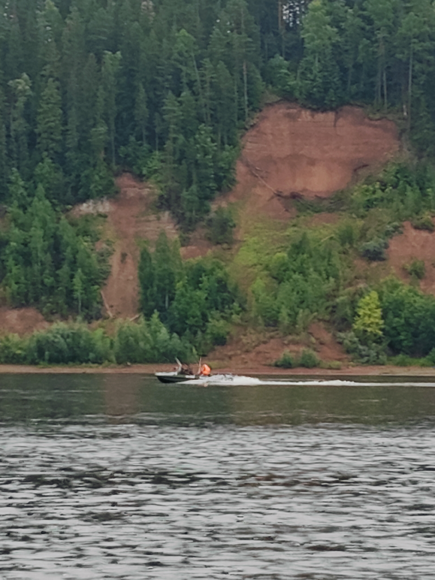 The image captures a tranquil scene of a boat navigating a wide river in Noviy, Russia. A small, dark motorboat, with two individuals onboard wearing life vests, is seen moving from left to right, leaving a white wake behind it. The boat and its occupants are the central focus of the activity.
The setting is a lush, natural landscape dominated by a steep, treelined embankment on the far side of the river. The embankment is characterized by exposed reddish-brown soil and a dense growth of green trees. The river itself appears calm, with ripples reflecting the overcast sky. The time of day seems to be late afternoon or early evening, judging by the soft lighting and muted colors. The weather appears to be cloudy, contributing to the serene atmosphere of the scene. No visible text is present in the image.