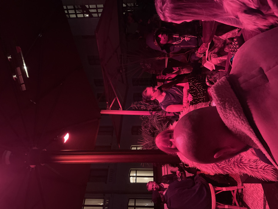 The image depicts a nighttime social gathering on what appears to be an outdoor or semi-outdoor terrace in Paris, France. The scene is illuminated predominantly by a strong, reddish ambient light. Overhead, large dark canopies or umbrellas are visible, supported by prominent poles, with visible heating elements suspended beneath. In the background, multi-story buildings consistent with Parisian Haussmannian architecture feature multiple illuminated windows. Sparse vegetation, including potted plants, is present.
Approximately ten individuals are discernible, engaged in social interaction. In the immediate foreground, a male with a bald head is observed from behind, wearing a brown jacket with a light-colored shearling collar. To his left, a partially obscured female with light-toned hair is wearing a shaggy faux fur coat. Further back on the left, a smiling male with short, light hair and a beard is seated, wearing a dark jacket. Central to the midground stands a female with dark hair pulled back, wearing a dark, sleeveless top and patterned bottoms, looking downwards. To her right, a seated male wears a dark hooded jacket, sunglasses, and multiple prominent necklaces, gesturing with his right hand. To the far right, a female with long, light hair is seen from behind, facing the gesturing male. Dark-colored tables are present with various items, including drinks, and a light-colored stool is visible on the left.