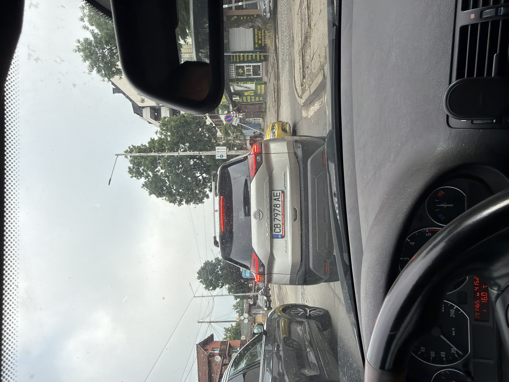 The image presents a daytime street view from inside a vehicle, looking through the windshield. The perspective is from the driver's seat, showing a portion of the dashboard, steering wheel, and rearview mirror. The weather appears overcast, with a bright, cloudy sky and some streaks on the windshield.
Ahead, a silver Nissan X-TRAIL e-POWER SUV is visible, seemingly stopped in traffic. Its rear license plate, "BG CB 7978 AE," identifies it as registered in Bulgaria, with "CB" indicating the Sofia region.
The dashboard of the observer's vehicle shows an odometer reading of 237,465 km and an outside temperature of 16.0 °C.
Beyond the Nissan, more cars are in line, including a yellow vehicle. The street is lined with buildings, some appearing to be shops, featuring signage in Cyrillic script. Visible text on storefronts includes "ПО-НИСКИ ЦЕНИ" (lower prices), "по-дълъг" (longer), "ГРАТИСЕН ПЕРИОД" (grace period), and "GSM" (mobile phones). A "no entry" road sign is also discernible. Trees and utility poles with overhead wires are present along the road, indicating an urban setting in Sofia, Bulgaria, likely during a traffic queue.
