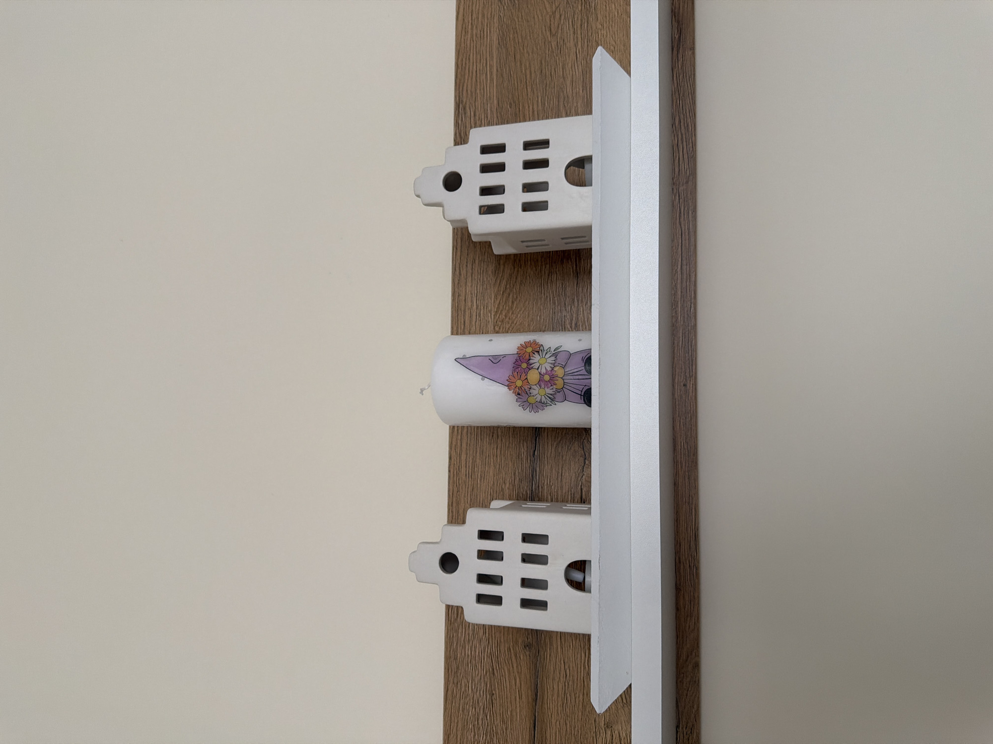 The image depicts a shelf mounted on a wall. On the shelf are two white ceramic decorative houses and a white cylindrical candle. The houses are shaped like Dutch gabled houses, with rectangular cutouts representing windows and doors. The candle has a graphic of a gnome holding a bouquet of flowers. The background wall is painted a light beige color, and the shelf is mounted against a wooden panel. The shelf appears to be a floating shelf. There is no indication of the city of Pazardzhik, Bulgaria.