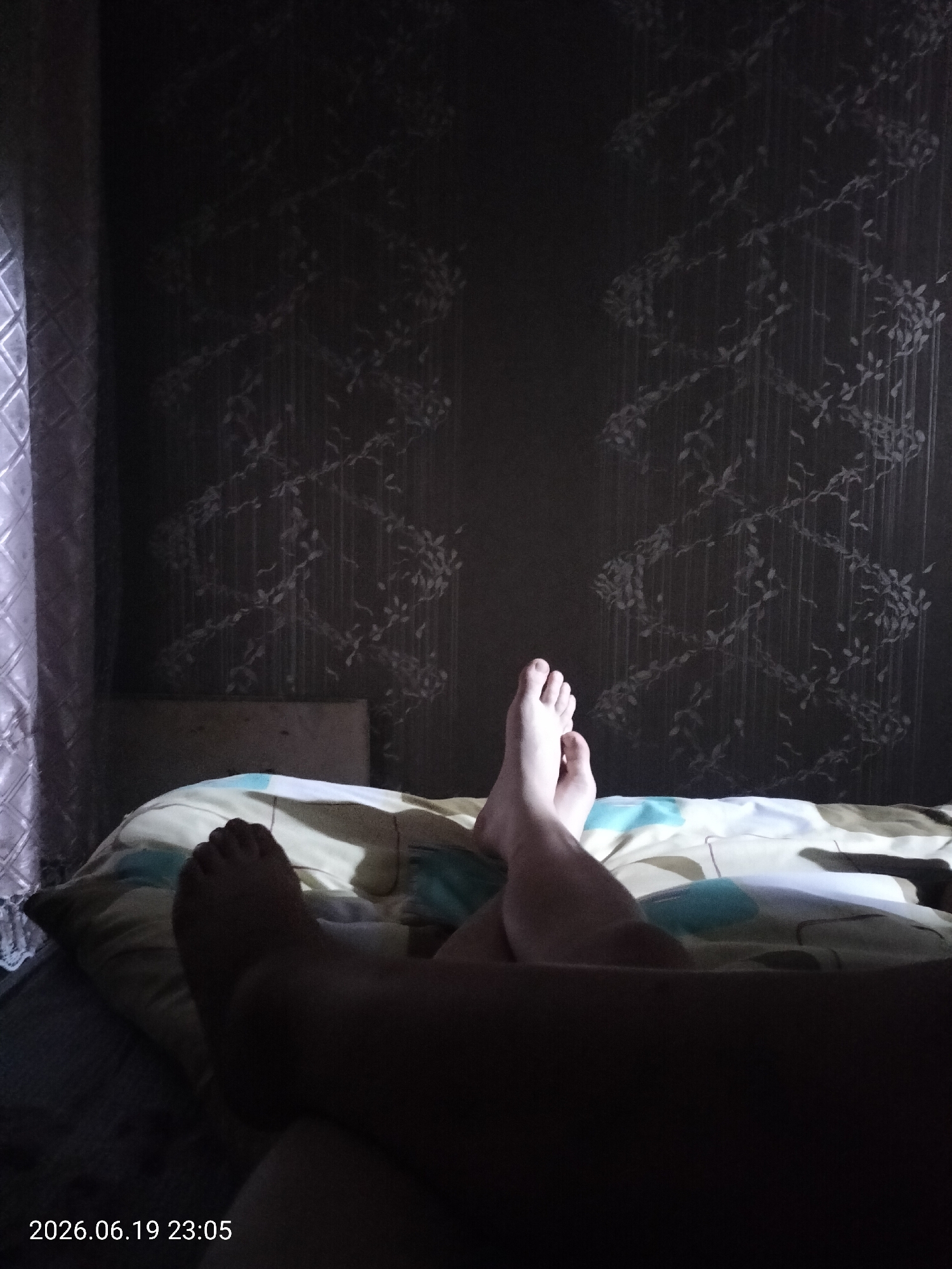The image shows a person's legs and feet resting on a bed. The person appears to be lying down, possibly in a bedroom, as indicated by the presence of a pillow and bedding. The background features wallpaper with a subtle floral or vine pattern. A curtain or window covering is visible on the left side of the frame. The lighting in the room is dim, suggesting it is nighttime or the lights are turned down. The timestamp in the bottom left corner indicates the date and time as 2026.06.19 23:05. The overall mood is relaxed and private. No specific weather conditions are discernible. The location context provided is Ridder, Kazakhstan.