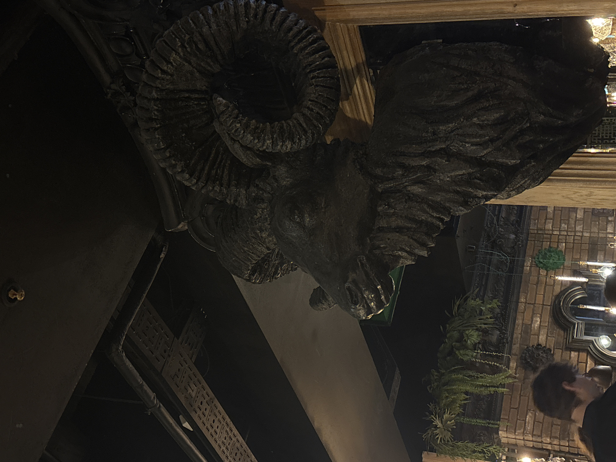 A dimly lit scene in Saint Petersburg, Russia, shows a dark, ornate ram's head sculpture mounted on a wall. The sculpture appears to be made of stone or a similar heavy material. The sculpture has curved horns and detailed features. Below the sculpture, there are metal fixtures, possibly pipes or conduit, running along the wall. A beam or ledge is present below the ram head. In the background, there is brickwork, greenery, and people. A man with dark hair is partially visible, facing the background brickwork and greenery. The environment suggests an interior location such as a bar. Chandeliers are visible.