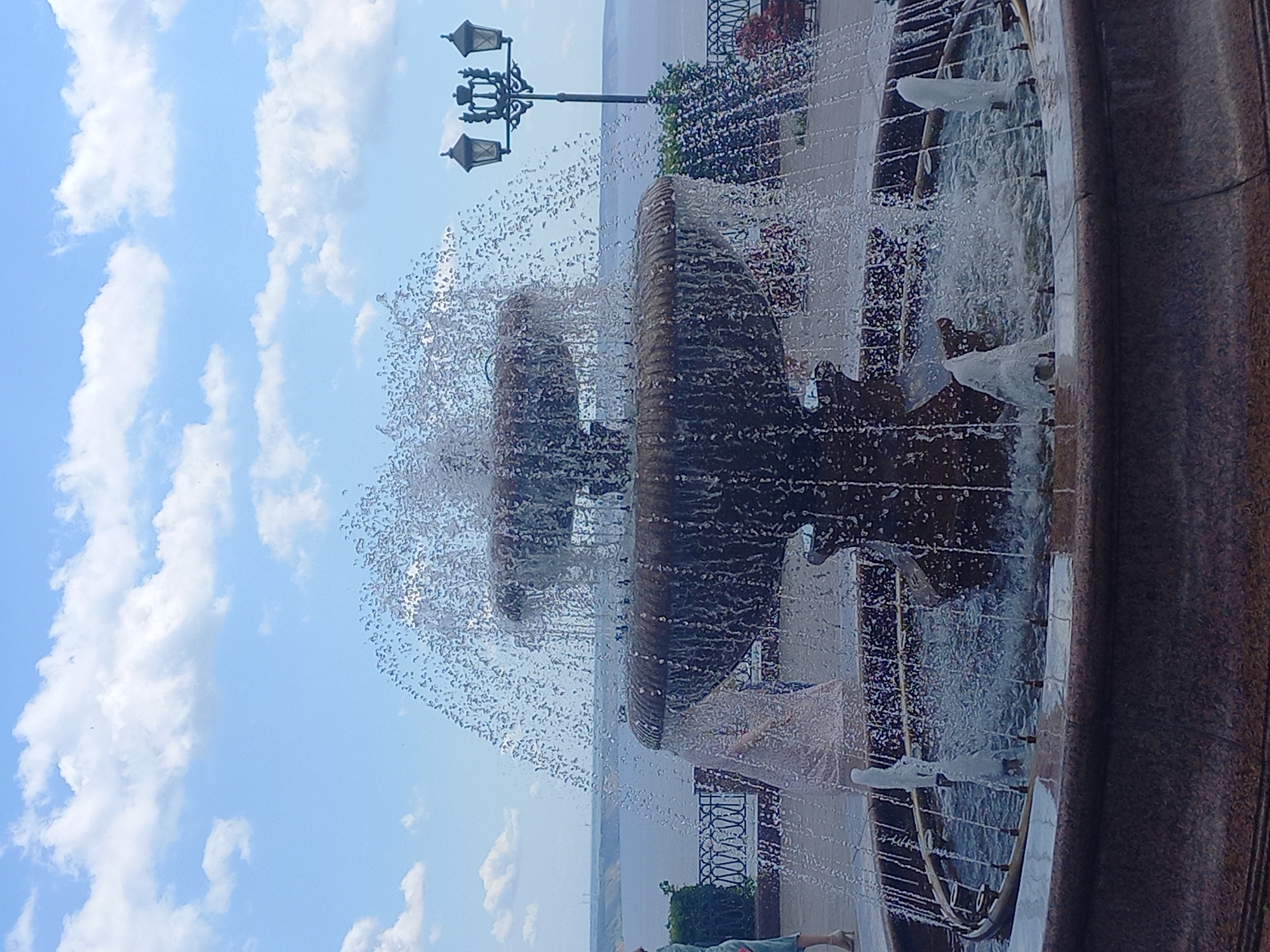 This is a photograph of a multi-tiered fountain in Ulyanovsk, Russia, situated on a promenade overlooking a body of water. The fountain is made of stone with a textured, weathered appearance and is actively spraying water, creating a dynamic and misty effect. Several jets of water are visible, some shooting upwards and others cascading down the tiers. In the background, a wide river or bay is visible, with a distant, hazy shoreline. A decorative lamppost with multiple lamps stands to the right of the fountain. Several people are present on the promenade, including a woman in a light-colored dress walking away from the viewer and another person on the far left wearing light-colored clothing. The sky is blue with scattered white clouds. The ground around the fountain is paved. The overall scene depicts a public recreational area on a bright day.