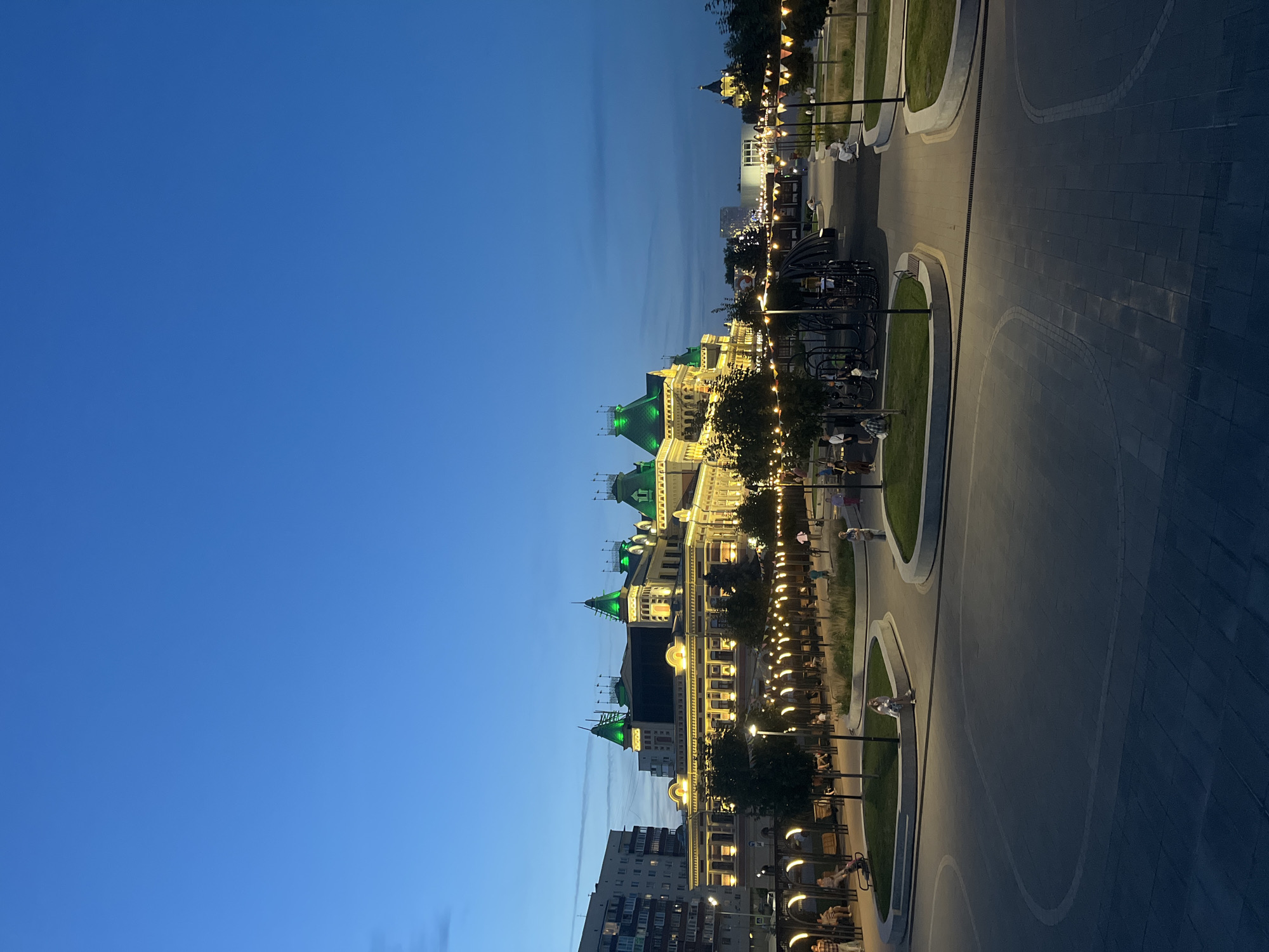 The image depicts a public square and a building in Nizhny Novgorod, Russia, at dusk. The building, illuminated with warm yellow lights, features green-capped towers and ornate architecture, possibly a historical landmark or a cultural institution. In the foreground, a paved plaza leads to a park area with grass, trees, benches, and string lights that create a festive atmosphere. Several individuals are visible, walking, sitting on benches, and interacting within the park, suggesting a casual, evening gathering. The sky is a deep blue, transitioning from twilight, with a few wispy clouds visible. To the left, a multi-story residential building stands, providing a contrast to the more decorative building. The overall scene conveys a sense of a lively urban evening in a well-maintained public space.