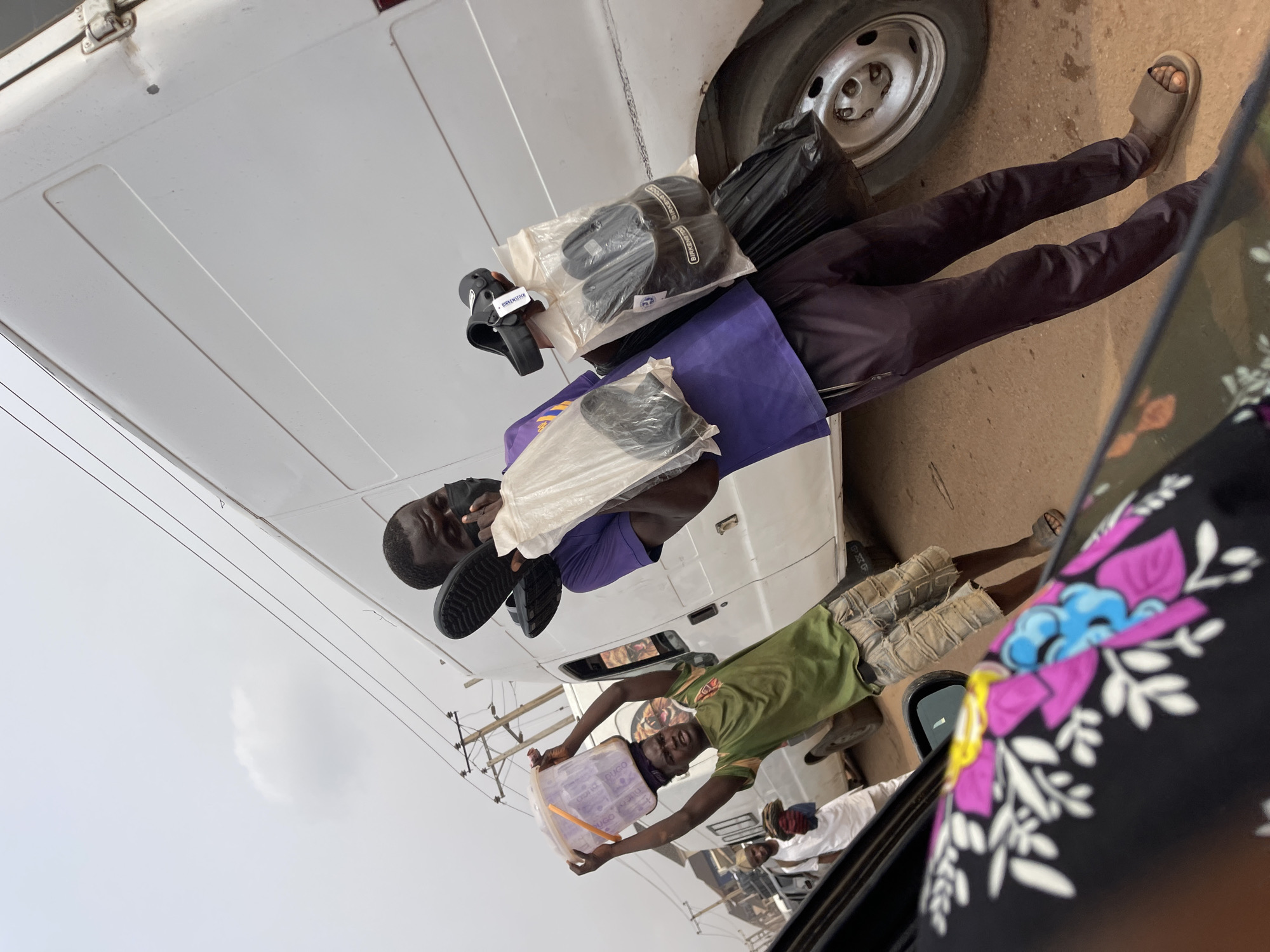 This image depicts a vibrant street vending scene in Ado, Nigeria, captured from the interior of a vehicle. The photograph is taken with a tilted perspective, looking out towards the street and other vehicles.
Two men are the primary subjects, both appearing to be hawking goods to passing or stationary vehicles. In the foreground, a man wearing a purple shirt and dark trousers holds up multiple pairs of black sandals or slides. Some of these are presented in clear plastic packaging, and the brand "Birkenstock" is clearly visible on their labels. A black plastic bag is also held near his arm.
Standing slightly behind and to the right, another man in a green t-shirt and patterned shorts holds up a large, clear plastic container filled with small, possibly individually packaged items, such as sachets of water or snacks. The word "UGO" is partially visible on the container. Both men are looking towards the camera, suggesting they are engaging with potential customers in the vehicle from which the photo was taken.
In the background, a white van or bus is visible, with parts of its wheel and side panel showing. Overhead power lines and utility poles can be seen against a bright, overcast sky. The bottom right corner of the image shows a black surface with a vivid floral pattern in pink, blue, and white, likely part of the car's interior from where the picture was shot. The scene occurs during daylight hours, under a cloudy sky, typical for urban roadside commerce.