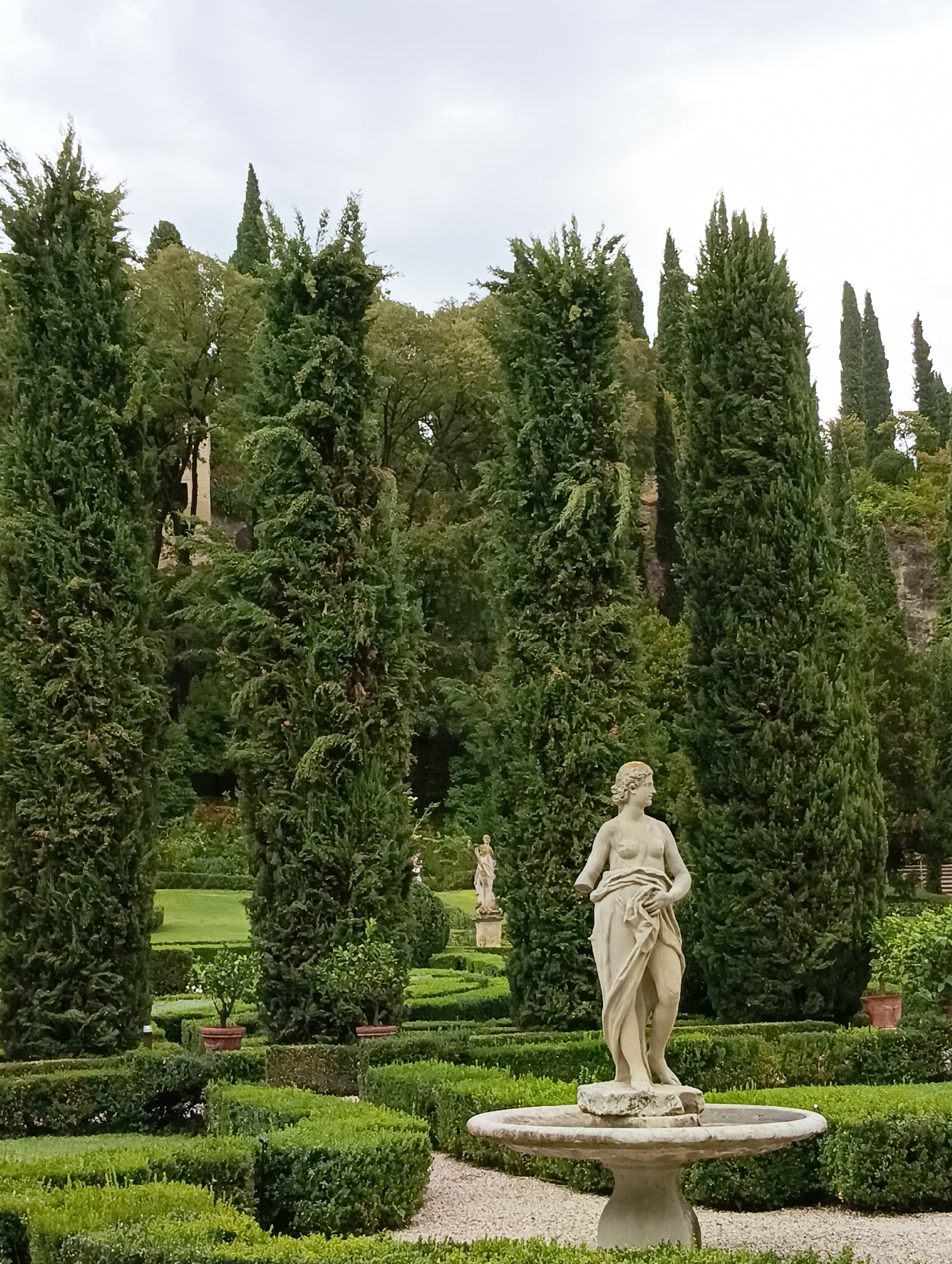 This image depicts the Giardino Giusti in Verona, Italy. The scene is a formal Italian garden featuring manicured boxwood hedges in geometric patterns. In the foreground, a classical stone statue of a partially draped female figure stands on a pedestal in the center of a circular fountain. Tall, slender cypress trees line the background, creating a sense of depth and grandeur. Beyond the cypress trees, a hillside covered in dense foliage and some visible stonework suggests a natural landscape. The sky is overcast, casting a soft, diffused light on the scene. Another statue is visible in the distance to the left, partially obscured by the cypress trees.